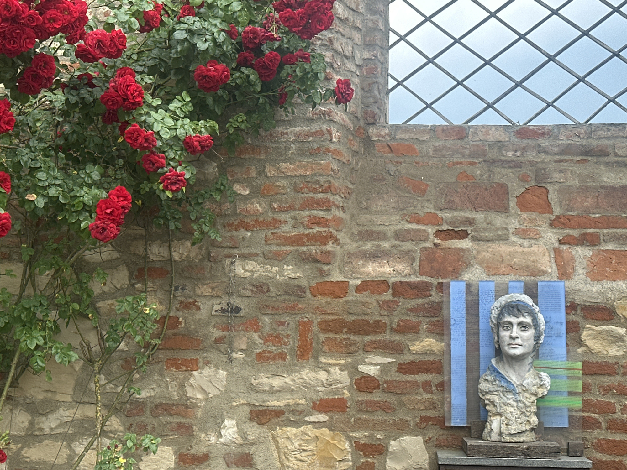 A bust of a woman is displayed against a brick wall. The bust is made of stone and is set on a wooden base. Behind the bust is a blue and white striped panel with green stripes on the right side. The brick wall is made of red and tan bricks. To the left of the bust, a rose bush with red roses is growing on the wall. A window with a diamond pattern is visible in the upper right corner.