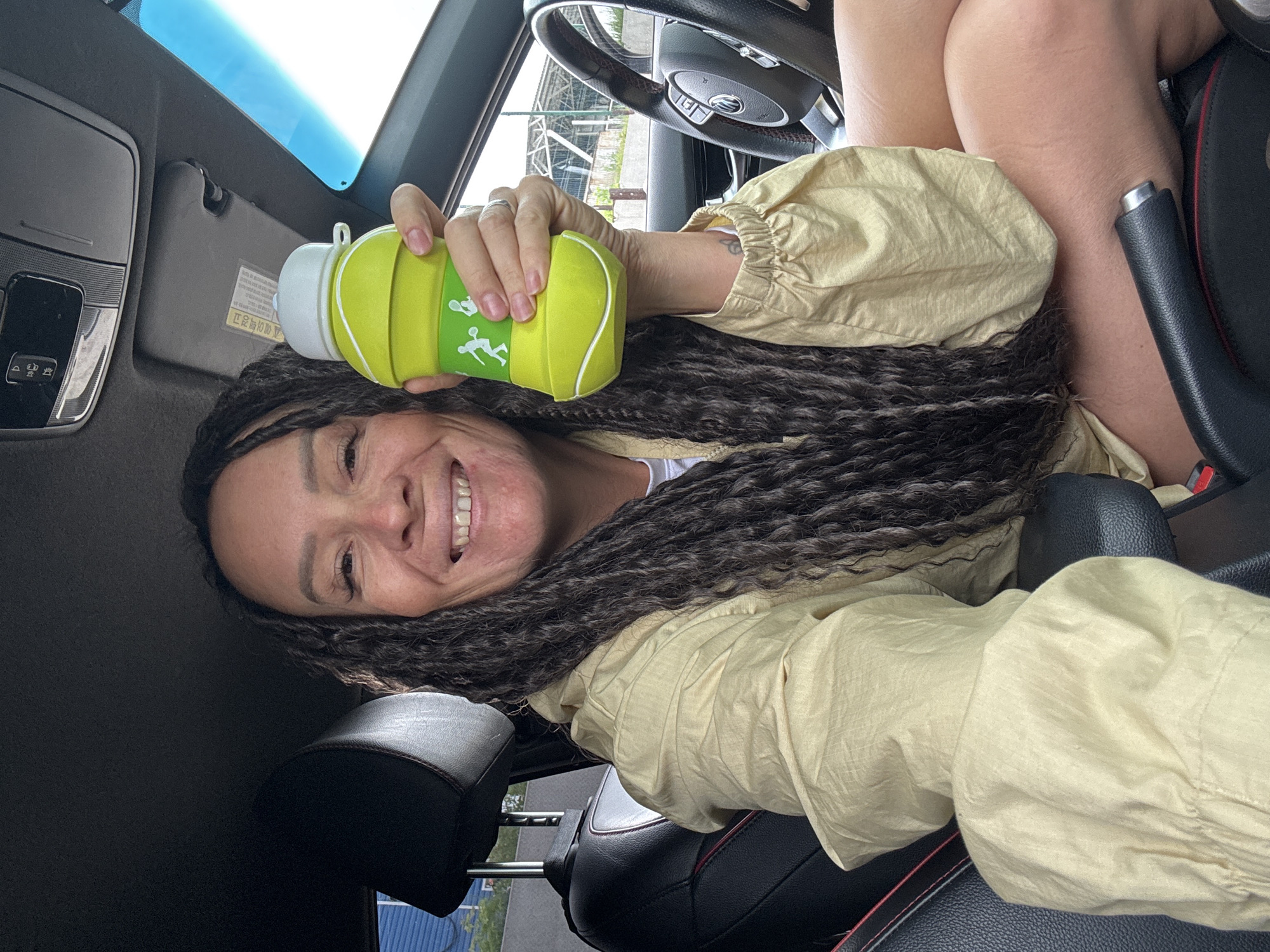 This rotated selfie captures a woman inside a car, smiling broadly at the camera. She has long, dark, braided hair and is wearing a light yellow or beige long-sleeved top or jacket. Her bare legs are visible in the lower right of the frame, positioned within the car's interior.
In her right hand, she holds a distinctive lime green water bottle, designed to resemble a tennis ball with white seam markings and a graphic of two stylized figures playing tennis. The car interior features black leather seats with red stitching, a portion of the steering wheel (suggesting a left-hand drive vehicle), and an overhead console with controls. A sun visor with some blurry, likely Cyrillic, text is also visible. Through the car's windows and sunroof, a bright, clear daytime sky and indistinct outdoor scenery are apparent.
The woman's wide smile conveys a cheerful and relaxed mood. The visible Cyrillic text on the sun visor and the left-hand drive car configuration align with the provided location context of Chaikovsky, Russia.