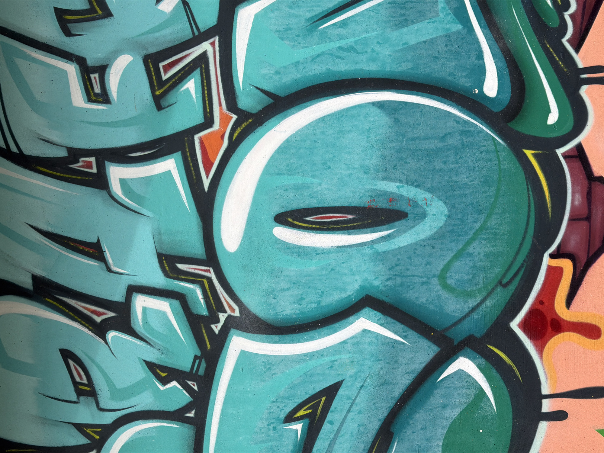Close-up of urban graffiti art. A predominant turquoise and teal palette with white and black outlining details dominates the left side. A large, stylized eye-like shape with a narrow horizontal dark center is centrally positioned, surrounded by a thick, white outline. Contrasting colors and patterns are visible on the right edge of the frame, including patches of pink, maroon, and green. The artwork contains sharp, geometric shapes and curves. The location is not discernible.