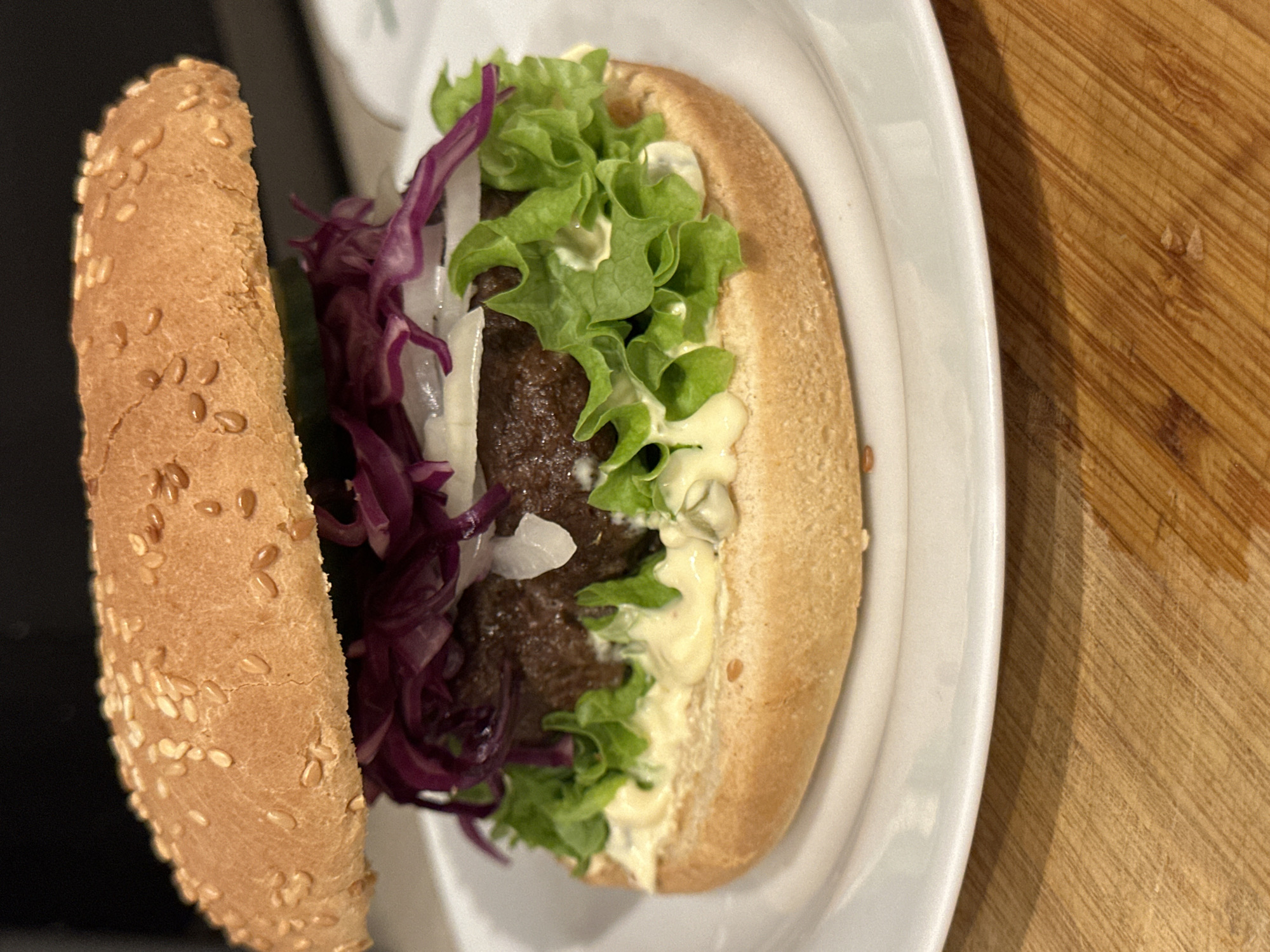 A close-up, eye-level shot shows a hamburger on a white plate. The hamburger is stacked with a beef patty, lettuce, sliced red cabbage, sliced white onion, and a slice of cucumber, all between a sesame seed bun. The hamburger is placed on a white plate which is on a wooden surface. The scene is set indoors. There are no people or animals visible in the image. This image is not sufficient to determine its location within Krefeld, Germany.