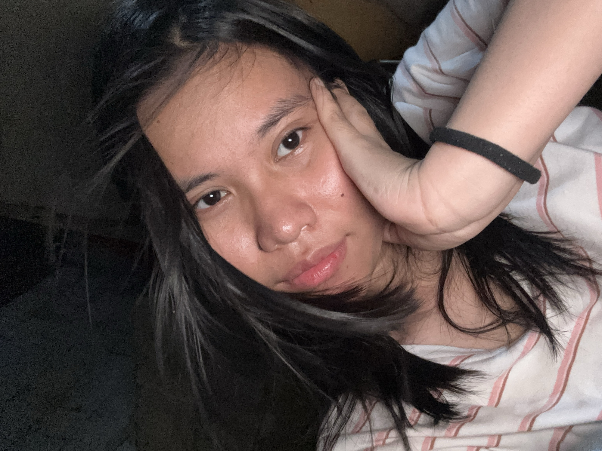 A close-up, low-angle selfie shows a young person with dark, shoulder-length hair resting their head on their right hand. The person is wearing a white t-shirt with thin pink and brown stripes. Their left eye is visible, and their mouth is slightly open. The background is dark and indistinct. The image was taken in East Jakarta, Indonesia.