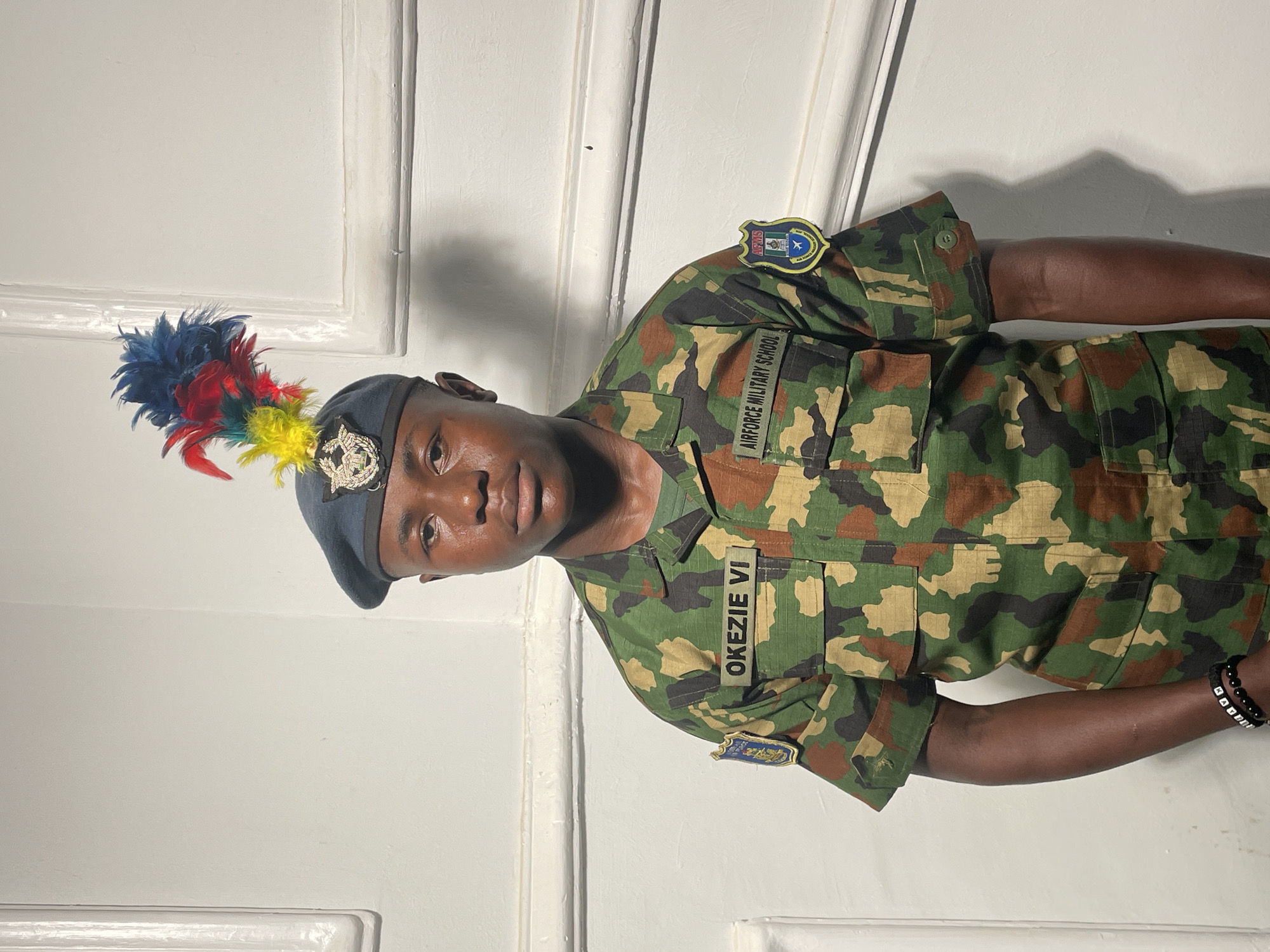 A man in an Airforce Military School uniform, labeled "OKEZIE VI," stands indoors against a white door. The uniform is camouflage pattern, with patches and insignia indicating affiliation with the Airforce Military School. He wears a beret adorned with a crest and colorful feathers. The photo appears to be taken in Barakin Akawo, Nigeria, based on the Airforce Military School affiliation. He also has a black beaded bracelet.