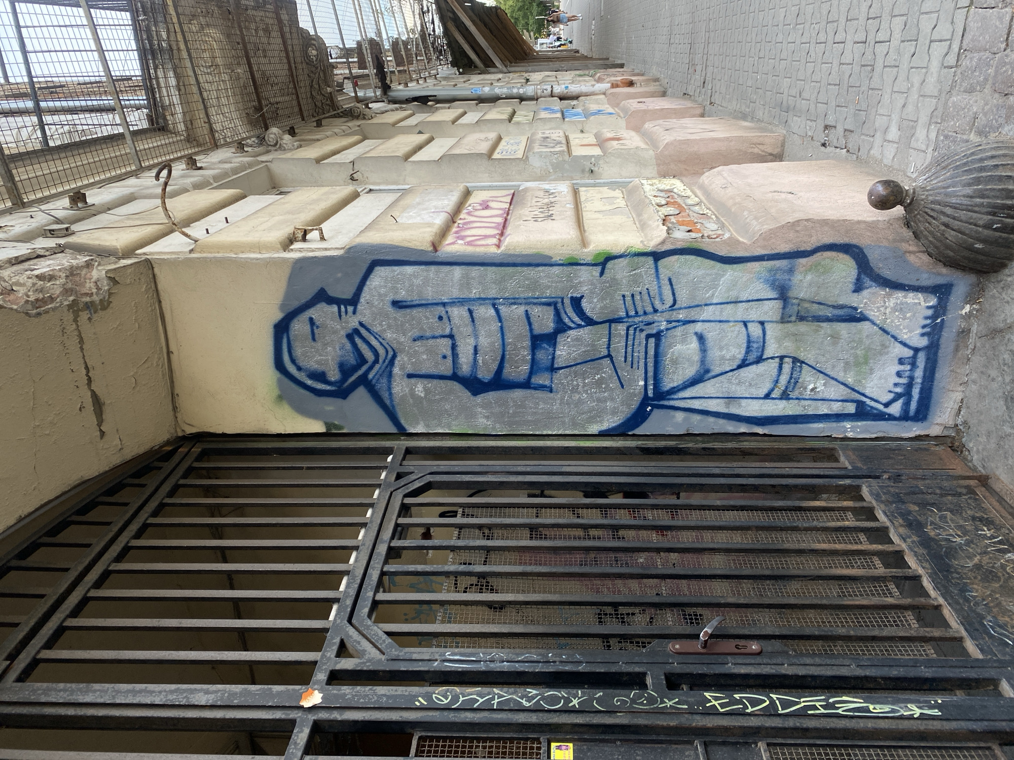 The image depicts a street scene in Warsaw, Poland, featuring a weathered building with a prominent metal gate on the left. The gate, constructed with vertical bars and a more robust frame, shows signs of age and rust, with some graffiti visible on its surface and behind it through the mesh. To the right of the gate, a building wall is adorned with graffiti, specifically a large blue outline of a humanoid figure with stylized facial features and segmented body. The building's facade displays crumbling plaster and architectural details, with a sidewalk of cobblestones extending along its length. Several people are visible in the distance walking along the sidewalk, and the overall impression is one of an urban environment with elements of decay and street art.