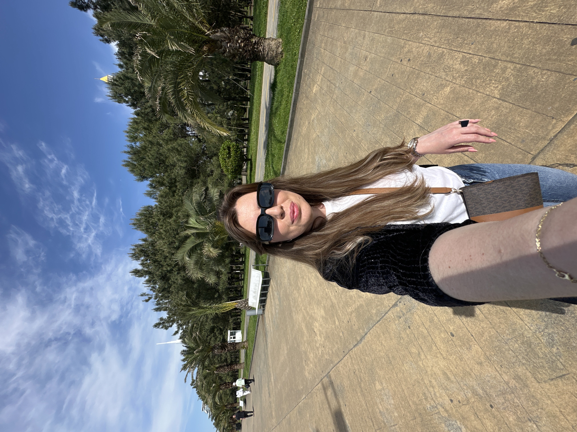 The image is a selfie of a woman, likely taken on a sunny day. The background shows a wide pedestrian walkway, probably in a park or public area. There are trees and palm trees lining the walkway, suggesting a subtropical or tropical climate. In the distance, a tower-like structure can be seen. Based on the style of the architecture and the presence of palm trees, the location is probably Batumi, Georgia. The woman is wearing sunglasses and holding the camera at arm's length. She has a purse slung across her shoulder. The scene is outdoors with good lighting.