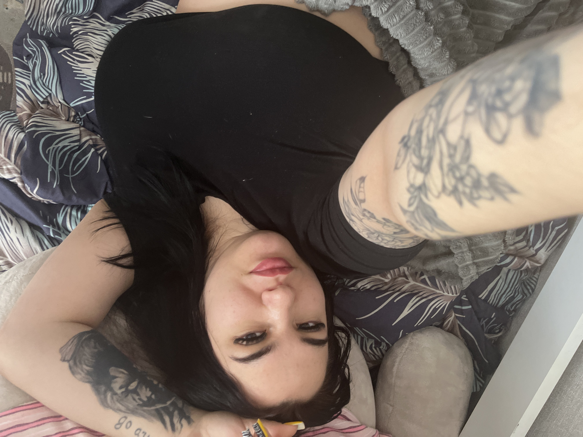 A woman lies on a bed with patterned bedding and a gray textured blanket. She wears a black shirt and has multiple tattoos on her arms. The tattoos are primarily black ink, and include floral designs and text. The woman's face is visible, featuring full lips, dark eyebrows, and dark hair. One hand rests near her face, displaying a ring and a manicured nail. The location is not specified in the image and cannot be determined from the provided visual information.