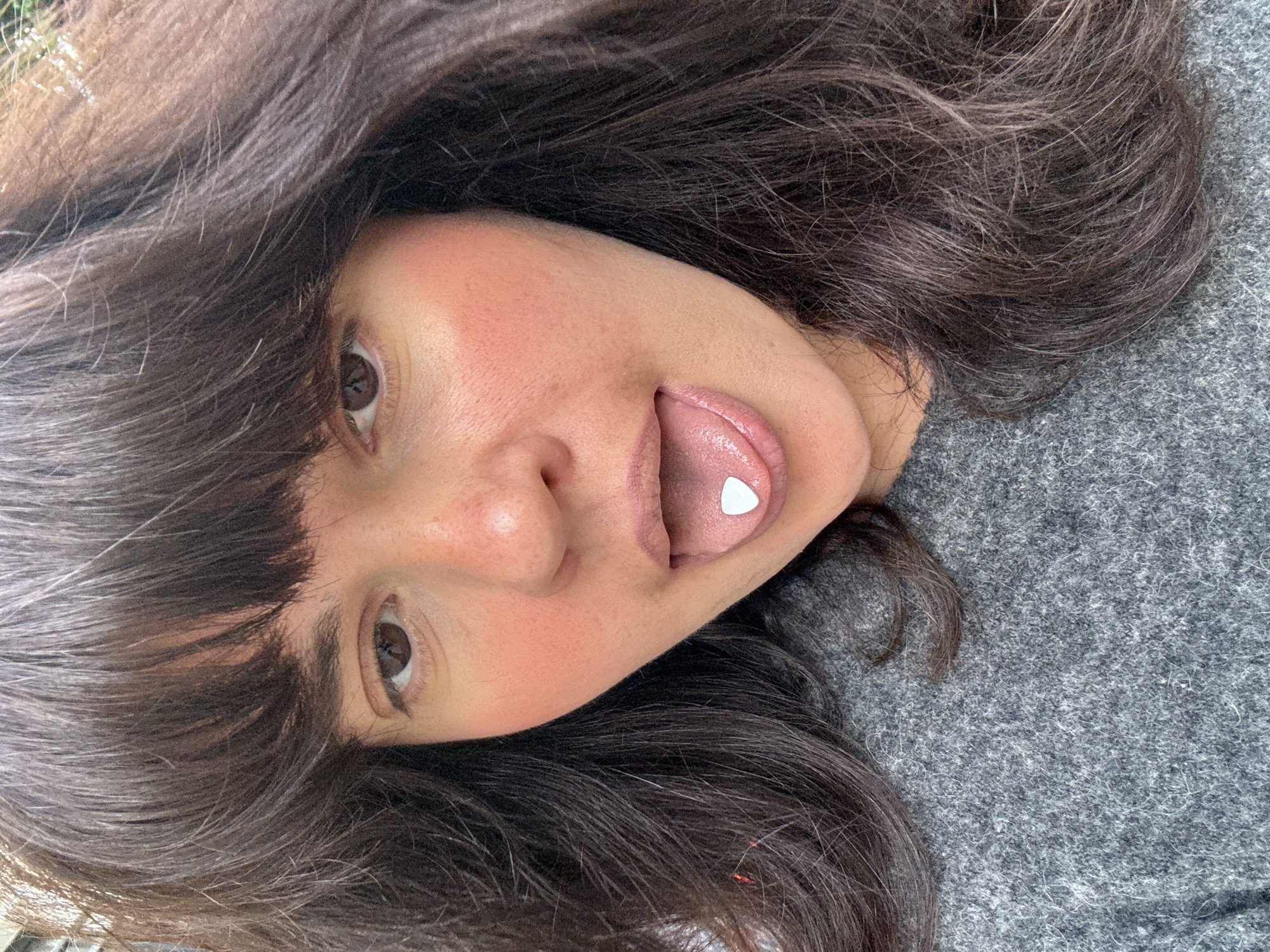 A woman with brown hair and brown eyes is lying on a gray fabric surface. Her head is tilted to the side, and her tongue is extended with a white, triangular object resting on it. Her face has rosy cheeks. The scene does not provide information to determine its location in Berlin, Germany.