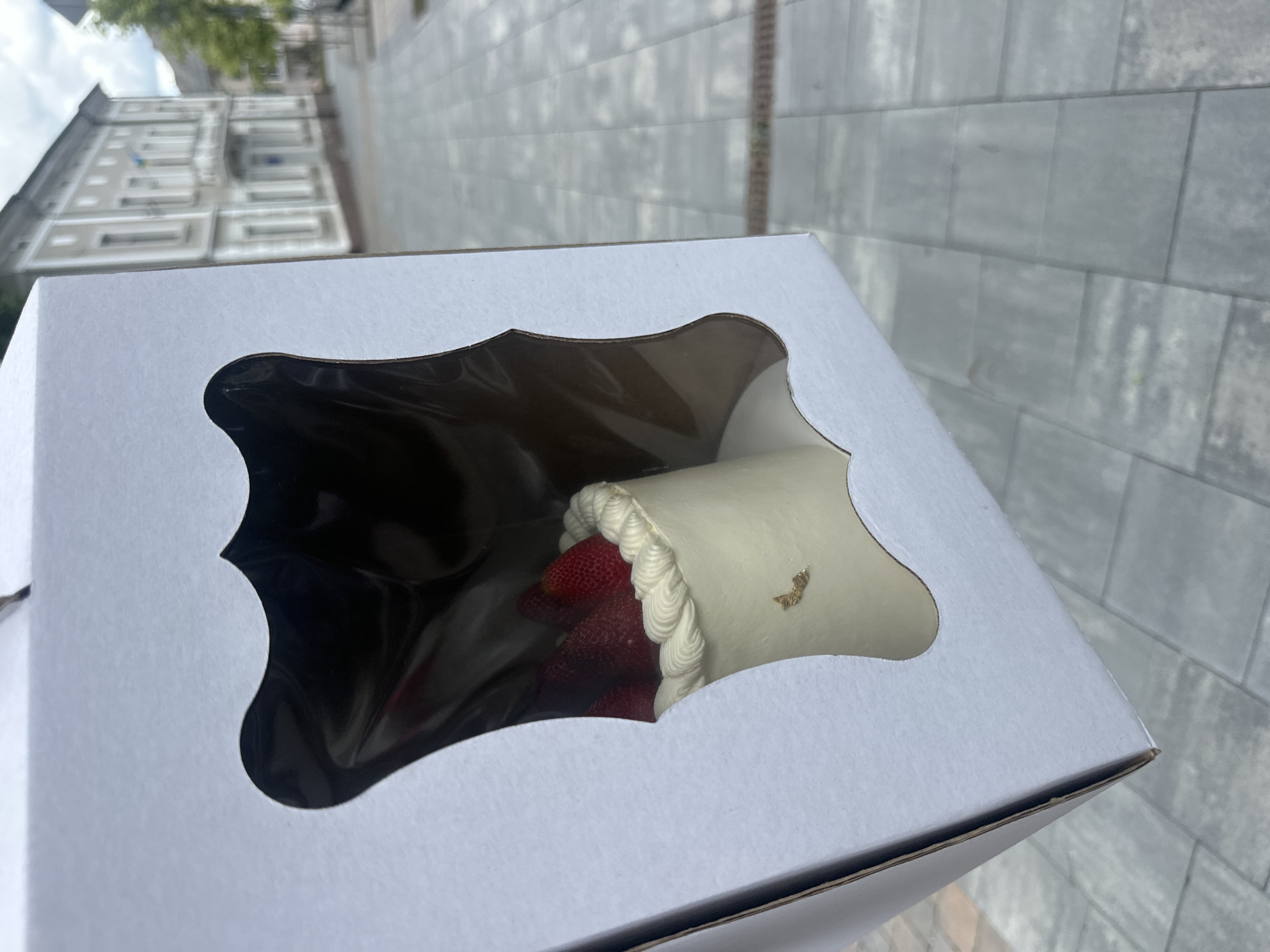The image shows a white cake box with a decorative cut-out revealing the contents: a cylindrical cake filled with strawberries and topped with white frosting. The cake is decorated with a small piece of gold leaf. The box is held at an angle, appearing to be outdoors. In the background, there is a paved area and a multi-story building. Based on the architecture of the building, the location is determined to be Ternopil, Ukraine.