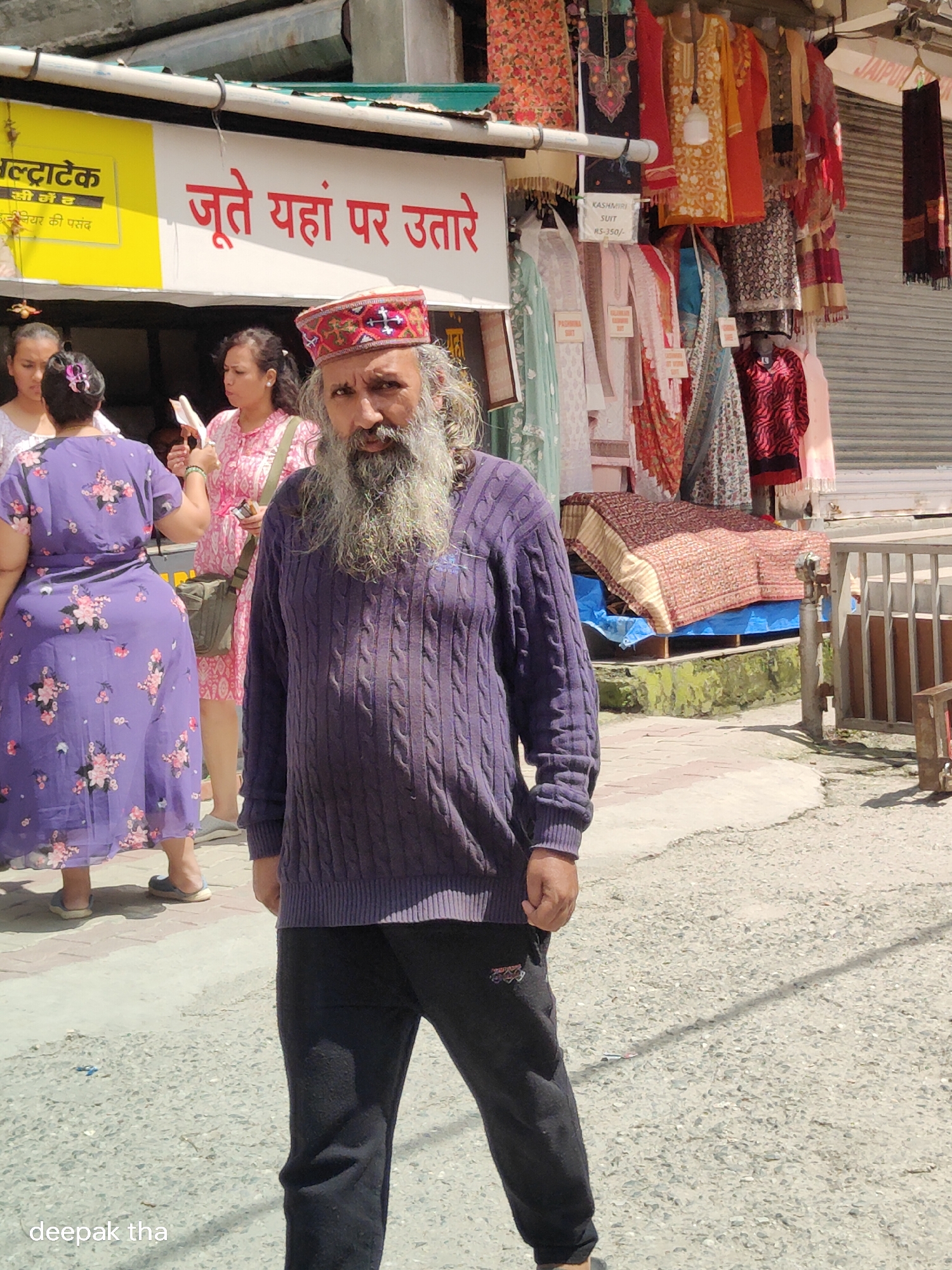 In Vashisht, India, a middle-aged man with a long grey beard and hair, wearing a purple cable-knit sweater and dark pants, stands prominently in the foreground. He has a traditional cap with red, white, and black accents on his head. Behind him, a row of shops displays various clothing items, including colorful women's suits and patterned fabrics hanging from racks. A yellow sign with Hindi text is visible, and further back, a woman in a purple floral dress and another in a pink patterned outfit are present. The ground is paved with a mix of asphalt and stone. The scene appears to be a street market on a sunny day.