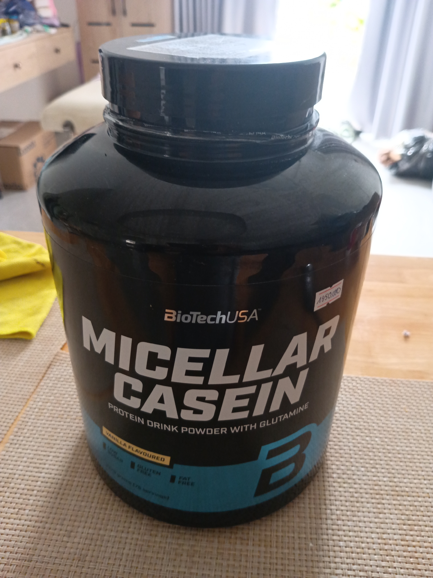 A large, black or dark blue plastic tub of BioTechUSA™ Micellar Casein protein powder sits on a light-colored woven placemat atop a wooden table. The container features the brand "BioTechUSA™" prominently, with "MICELLAR CASEIN" written in large white capital letters below it, followed by "PROTEIN DRINK POWDER WITH GLUTAMINE". A yellow band near the bottom indicates "VANILLA FLAVOURED", and smaller icons state "GLUTEN FREE" and "FAT FREE". A white label with red text, partially obscured, is affixed to the side, displaying "1950.000", likely indicating a price in Vietnamese Dong given the provided location context of Mũi Né, Vietnam. To the left of the tub, a bright yellow cloth is visible. The background shows an indoor setting with natural light coming from a window with white curtains. Light-colored wooden furniture, possibly a dresser or cupboard, and a chair are visible further back. Some dark bags or items are also present near the window. The image captures a static product shot in what appears to be a residential or hotel room during the daytime.