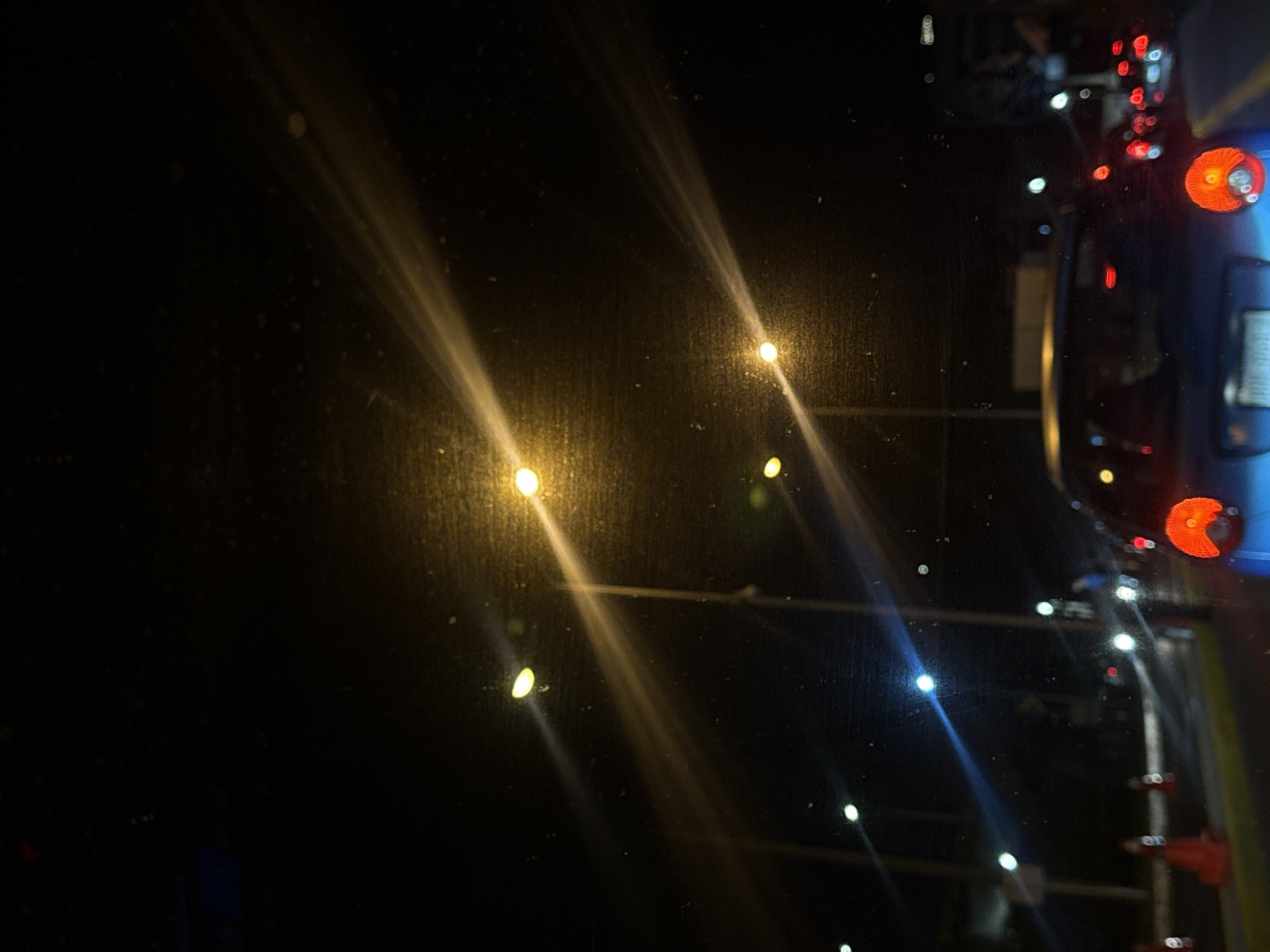 The image depicts a nighttime scene in an urban environment. Streetlights cast long, blurred streaks across a rain-streaked window. To the right, traffic lights and the tail lights of vehicles are visible, indicating the presence of a road. The scene appears to be viewed from inside a building or vehicle. The city and specific location are Unknown.