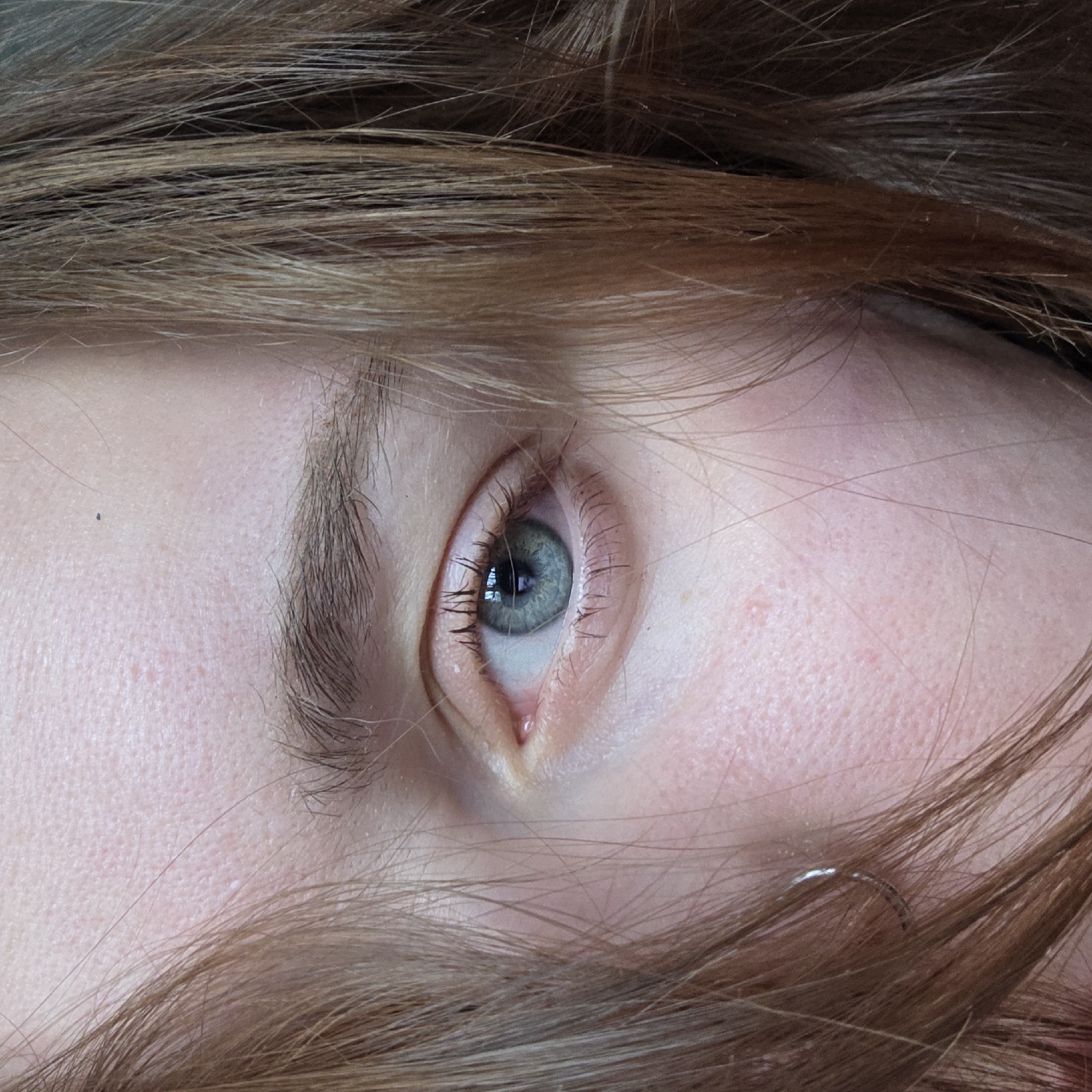 This is a close-up shot of a person's eye and surrounding facial area. The eye has a light blue-green iris and is framed by eyelashes. The person has light brown hair, some of which obscures parts of the face. The skin tone is fair. There is no indication of location in the city of Unknown, Unknown.