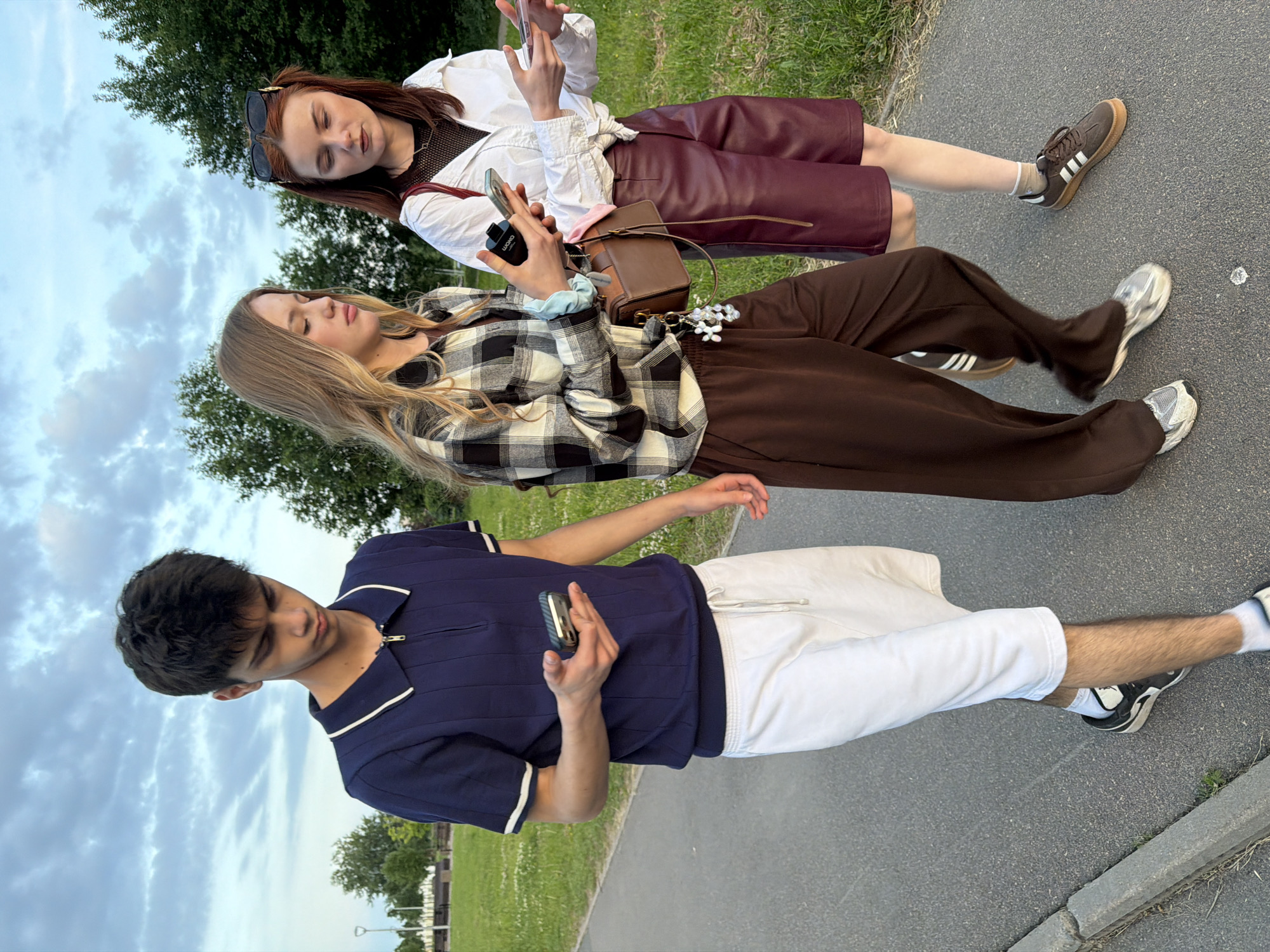 Three young people are walking on a paved path in a park-like setting. The person on the left, a male with dark hair, is wearing a navy blue collared shirt with white trim and white shorts. He is holding a phone in his right hand. In the middle, a female with blonde hair is wearing a black and white plaid shirt over a dark top and brown wide-leg pants. She is holding a phone in her left hand and has a brown crossbody bag with charms hanging from it. On the right, a female with reddish-brown hair, wearing sunglasses on her head, is wearing a white shirt and burgundy shorts. She is holding a phone in her right hand and is wearing white socks with brown sneakers. The background consists of green grass, trees, and a cloudy sky. The overall scene appears to be a casual outdoor excursion.