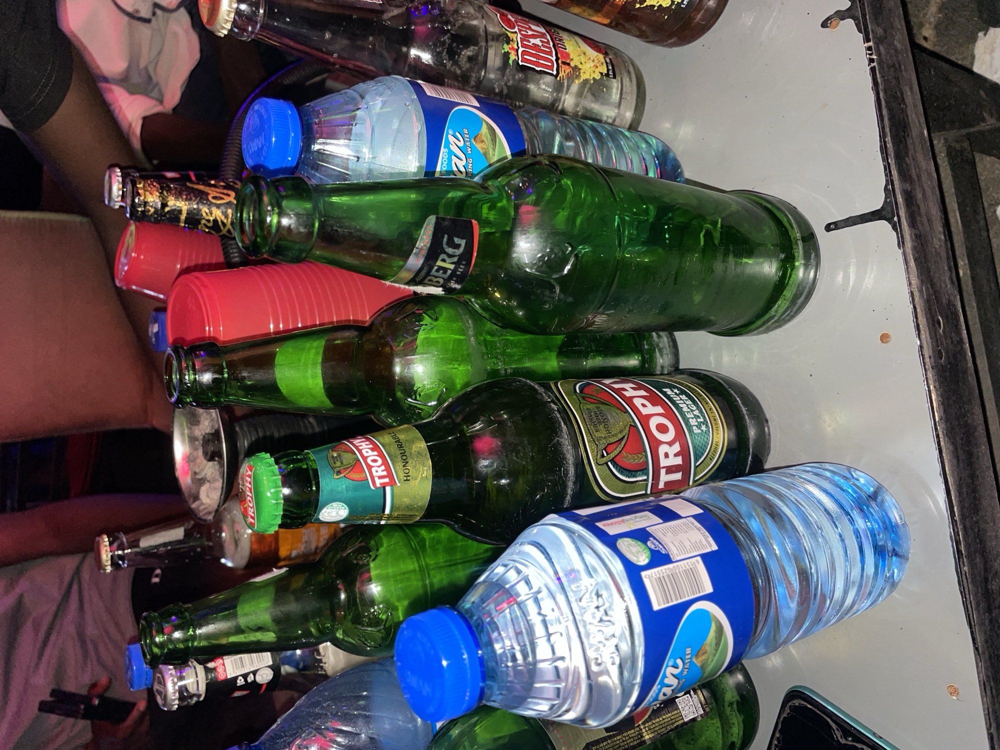 The image shows a collection of bottles and cups on a table. There are several green glass beer bottles, including brands such as "Trophy" and "BERG." There is also a bottle of "Dews" drink and several plastic water bottles with blue caps. Additionally, there are a couple of red plastic cups. The bottles and cups appear to be a mixed collection after a gathering. It is not possible to ascertain if the location is in Tumfure, Nigeria without more information.