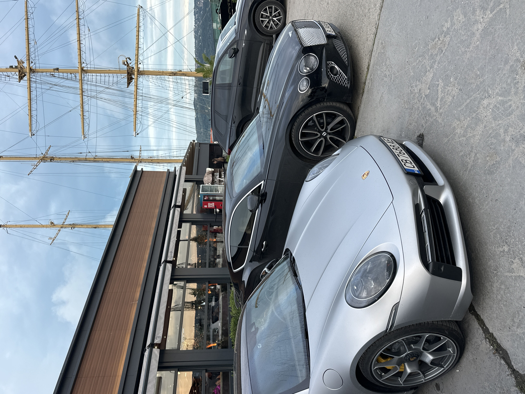 An outdoor scene captured in Varna, Bulgaria, depicts a luxurious waterfront setting. In the immediate foreground, a silver Porsche sports car, likely a 911 model, is prominently parked on a concrete surface. Its distinctive headlights, wheel design with yellow brake calipers, and a Bulgarian license plate "CB 7888 HE" are clearly visible.
Behind the Porsche, a black Bentley luxury car is parked, showcasing its iconic grille and circular headlights. Part of its Bulgarian license plate, starting with "B 382", can be seen. Further back, a dark-colored SUV, possibly another high-end vehicle, is partially visible. The presence of 'CB' and 'B' on the license plates indicates registration in Sofia, though the scene is set in Varna.
To the left of the parked cars, a modern building with large glass windows and dark frames stretches along what appears to be a promenade or dockside. A section of the building features brown cladding. Reflections in the windows hint at the sky and surrounding environment.
Dominating the background above the building and cars are the towering masts and intricate rigging of a large sailing ship, or tall ship, suggesting it is docked nearby. The ship stands against a bright, partly cloudy sky, with distant hills or land visible on the horizon. The overall lighting suggests it is daytime, possibly under an overcast sky or during late afternoon. There are no clearly visible people engaged in specific activities, and the scene appears calm. Some small, unidentifiable text can be seen on awnings on the building.