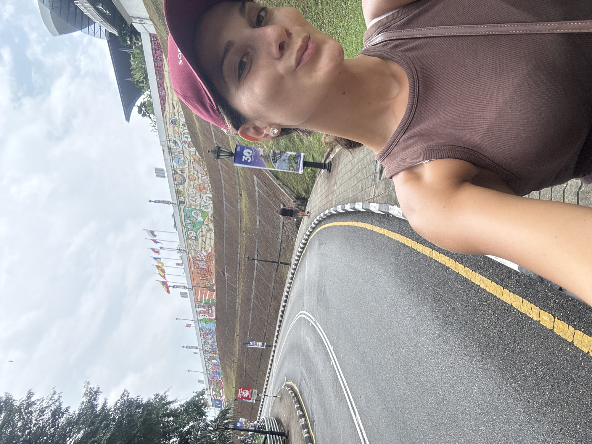 A medium shot shows a woman in a maroon baseball cap and brown sleeveless shirt, possibly a tank top. She is wearing a small, round earring. The woman seems to be taking a selfie.
In the background, on an inclined surface, is a very long and detailed mural. Several national flags are visible above the mural. A road with white and yellow painted lines is in the foreground, and streetlights are visible along the side of the road. Some trees are in the lower left corner.
The location is Kuala Lumpur, Malaysia, where the mural depicts the city's history.