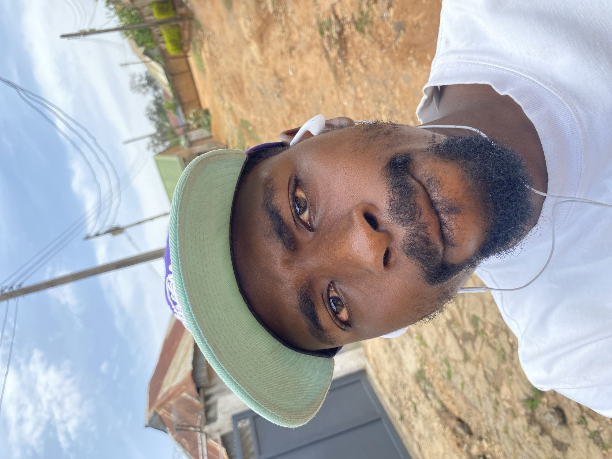 This close-up portrait, rotated 90 degrees clockwise, features a man with dark skin, prominent dark eyes, a full beard, and a mustache. He is looking directly at the camera with a neutral expression. He wears a white short-sleeved t-shirt and a light green baseball cap with a purple detail on the side. White wired earbuds are visible in his ears, with the wire extending down across his chest.
The background indicates an outdoor, urban or semi-urban setting under bright daylight in Jos, Nigeria. A clear blue sky with some scattered white clouds is visible, along with utility poles and power lines. Portions of sandy or dirt-covered ground with sparse greenery, blurred buildings, and a dark grey metal gate or door can also be seen in the background. There is no visible text in the image.
