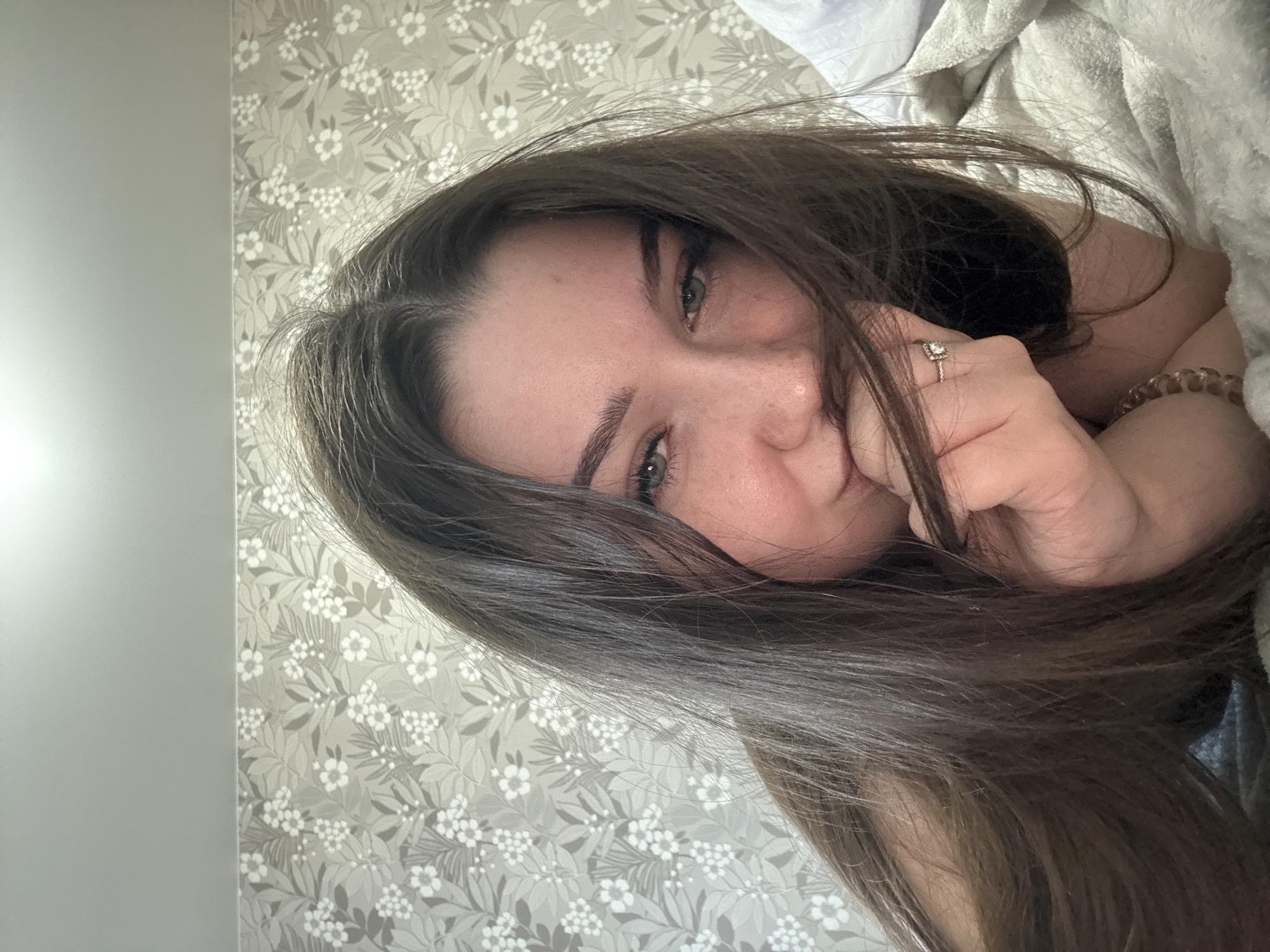 A medium shot shows a young woman with long, dark brown hair lying on a bed. She is looking at the camera with green eyes and her face is partially obscured by her hair. She is holding her hand to her mouth, and a ring with a square stone is visible on her right ring finger. She is wearing a bracelet on her left wrist. The background consists of a floral patterned wallpaper and a white blanket. The scene appears to be indoors, likely in a bedroom, in Zarechnaya (Gamovskoe S/P), Russia.