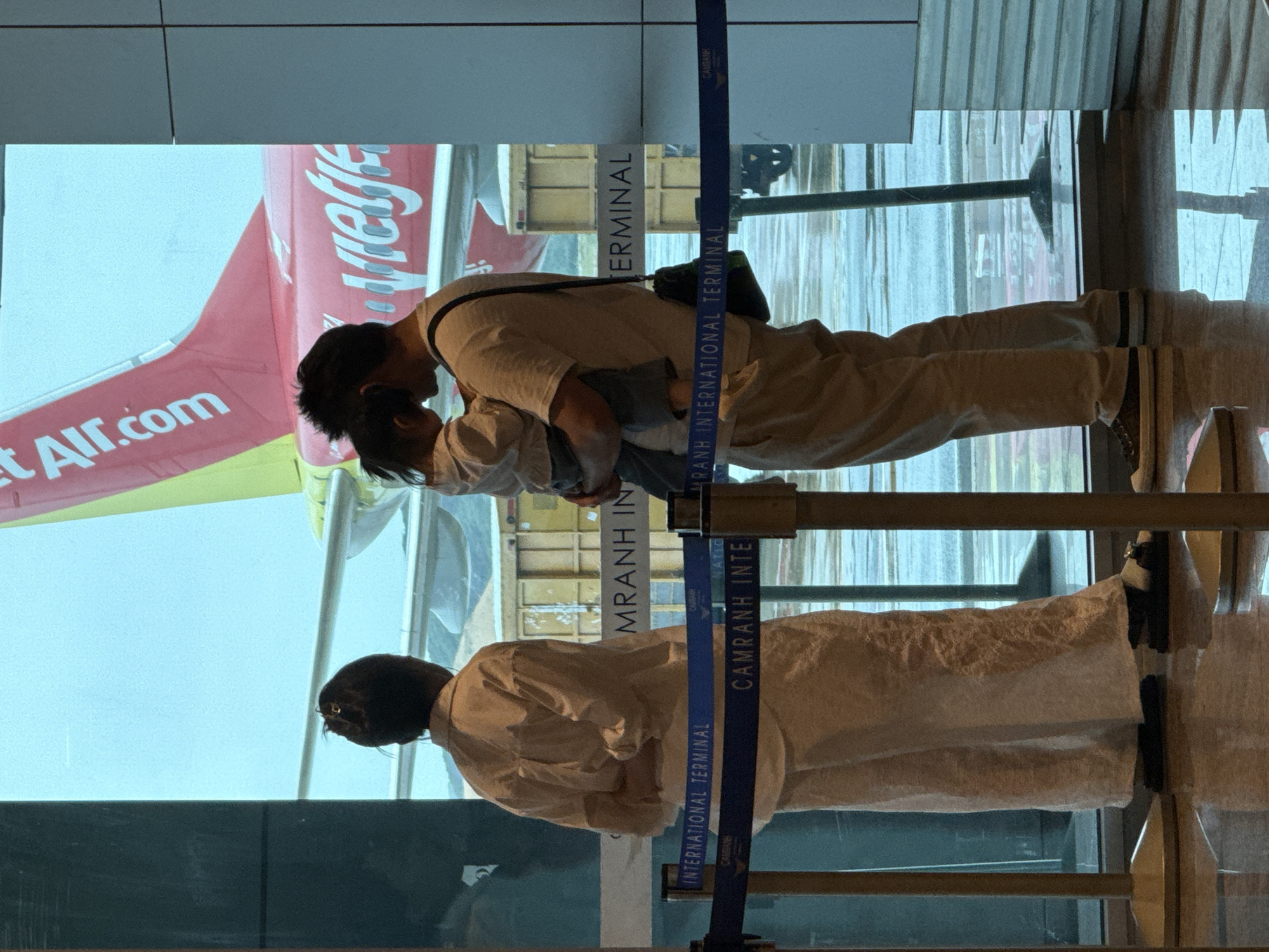 The image depicts a family at an airport gate in Cam Ranh, Vietnam, with an airplane visible outside. A woman stands to the left, facing away from the camera, wearing a light-colored, loose-fitting outfit. To her right, a man holds a young child, also facing away from the camera. The man is dressed in light-colored pants and a t-shirt, with a dark bag slung over his shoulder. The child is leaning into the man. In the background, a large red airplane with "VietJet Air.com" visible on its tail is parked on the tarmac. The scene is viewed through a large window, and blue stanchion ropes with "CAMRANH INTERNATIONAL TERMINAL" printed on them are in the foreground, indicating the location within the airport. The floor is reflective, showing the environment above.