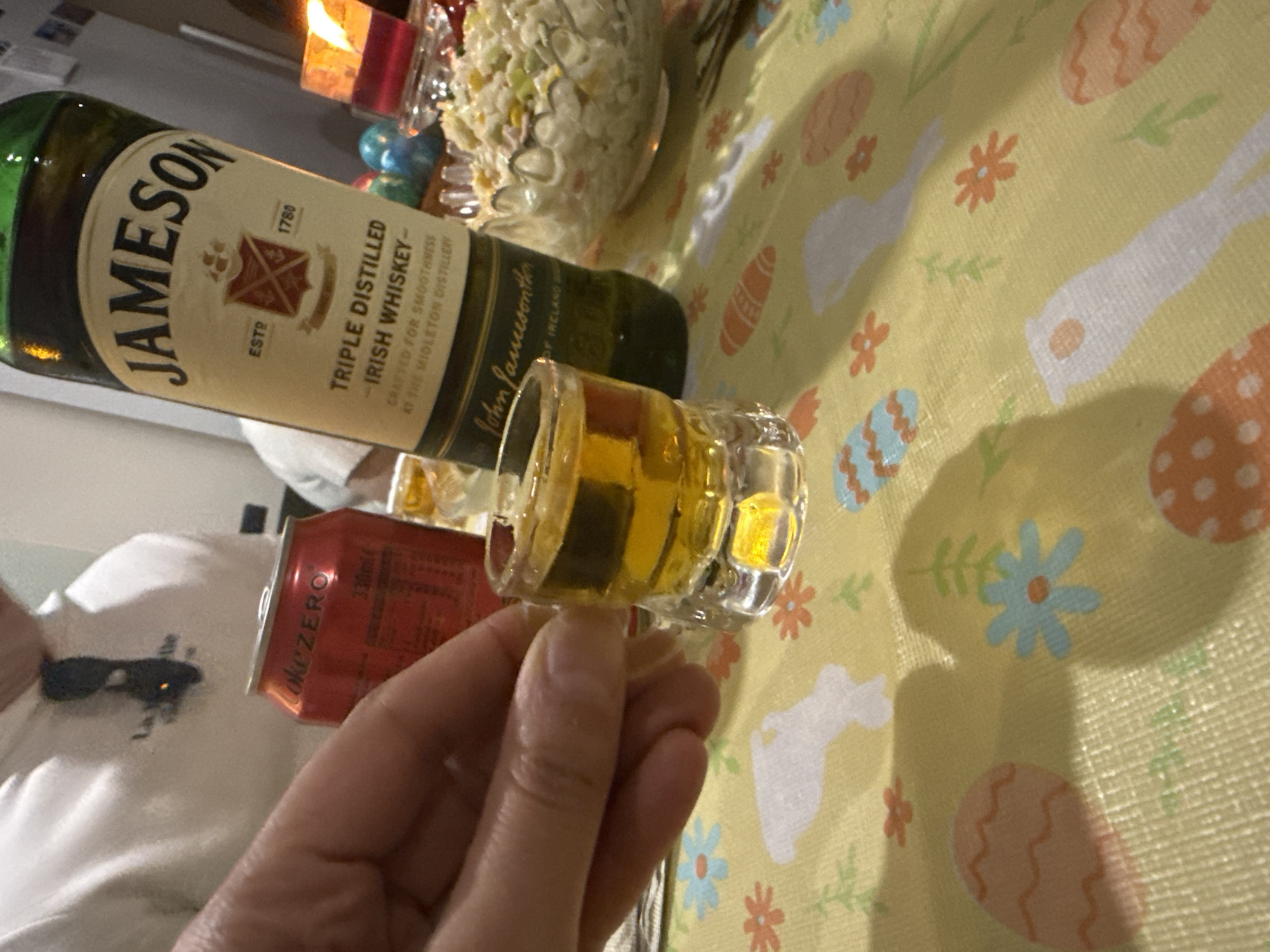 A person holds a shot glass filled with whiskey. A bottle of Jameson Irish Whiskey sits on a table covered with an Easter-themed tablecloth. A can of Coke Zero is visible, as is a bowl of food, possibly potato salad. The location in Gorleston-on-Sea, United Kingdom is unknown.
