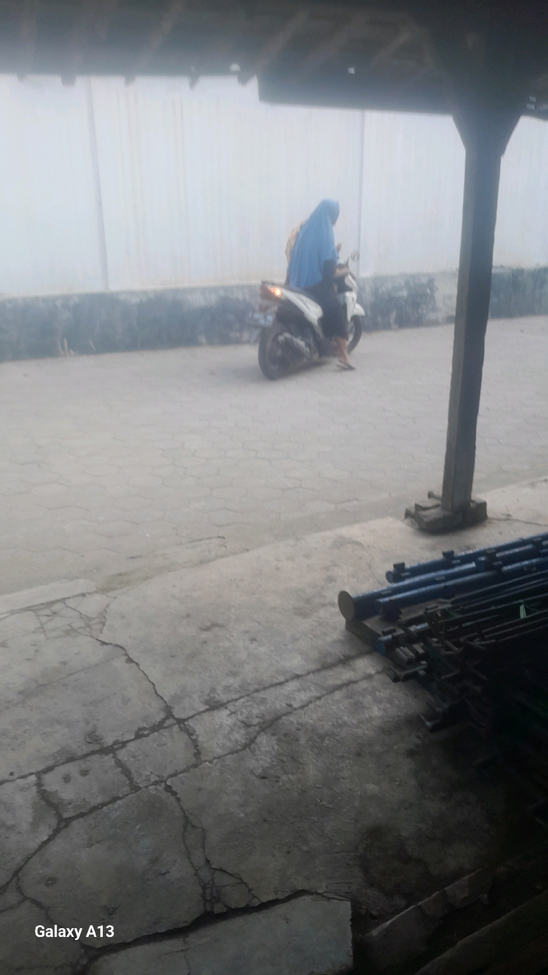 An outdoor scene, possibly in Ngelo, Indonesia, captured on a foggy or hazy day, shows a person wearing a blue hijab riding a white scooter. The person is seen from the side, and the scooter is moving on a paved surface made of interlocking stones. In the background, a light-colored wall stretches horizontally. In the foreground, a pile of blue metal pipes is stacked on the ground. The ground itself is a mix of cracked concrete and the paved interlocking stones. A dark, vertical pillar is visible on the right side of the image. The overall lighting is dim, suggesting overcast conditions or early morning/late evening light. The image has a low resolution and is somewhat blurry.