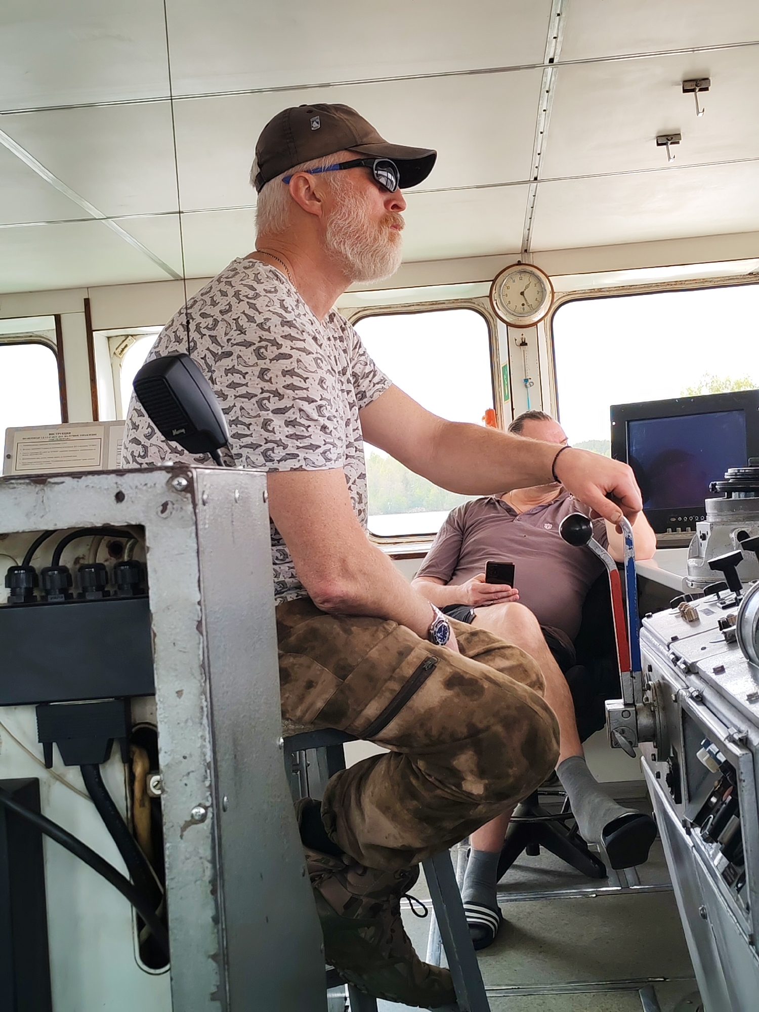 The image shows the interior of a boat, specifically the bridge or control room. A man wearing a patterned t-shirt, camouflage pants, sunglasses, and a baseball cap sits on a stool, operating controls. He has a white beard. Another man sits in the background, holding a cell phone, and wearing a short-sleeved shirt, shorts, socks, and sandals. A clock is visible on the wall, and windows show a river or lake environment. The location in the city of Yalosar', Russia cannot be determined from the image.
