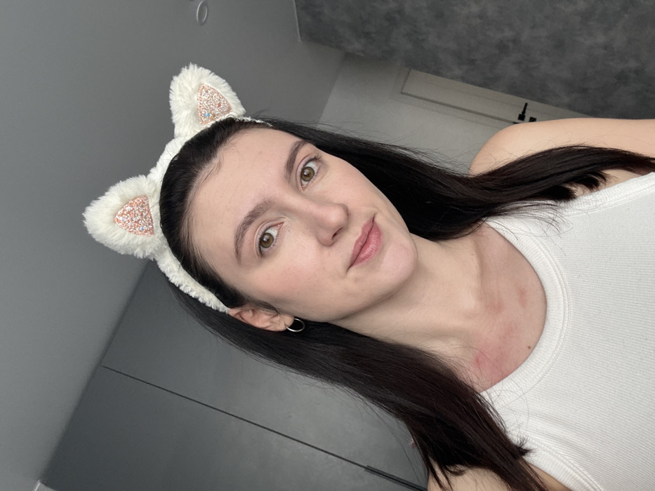 A woman with dark, straight hair and light skin wears a white tank top and a fuzzy white headband with cat ears adorned with sparkling accents. Her face is angled towards the camera, revealing her light brown eyes and a subtle smile. She has a piercing in her ear. The background consists of a gray wall and a glimpse of a white door or cabinet. The photo is taken in a well-lit environment, highlighting the details of her face and accessories.