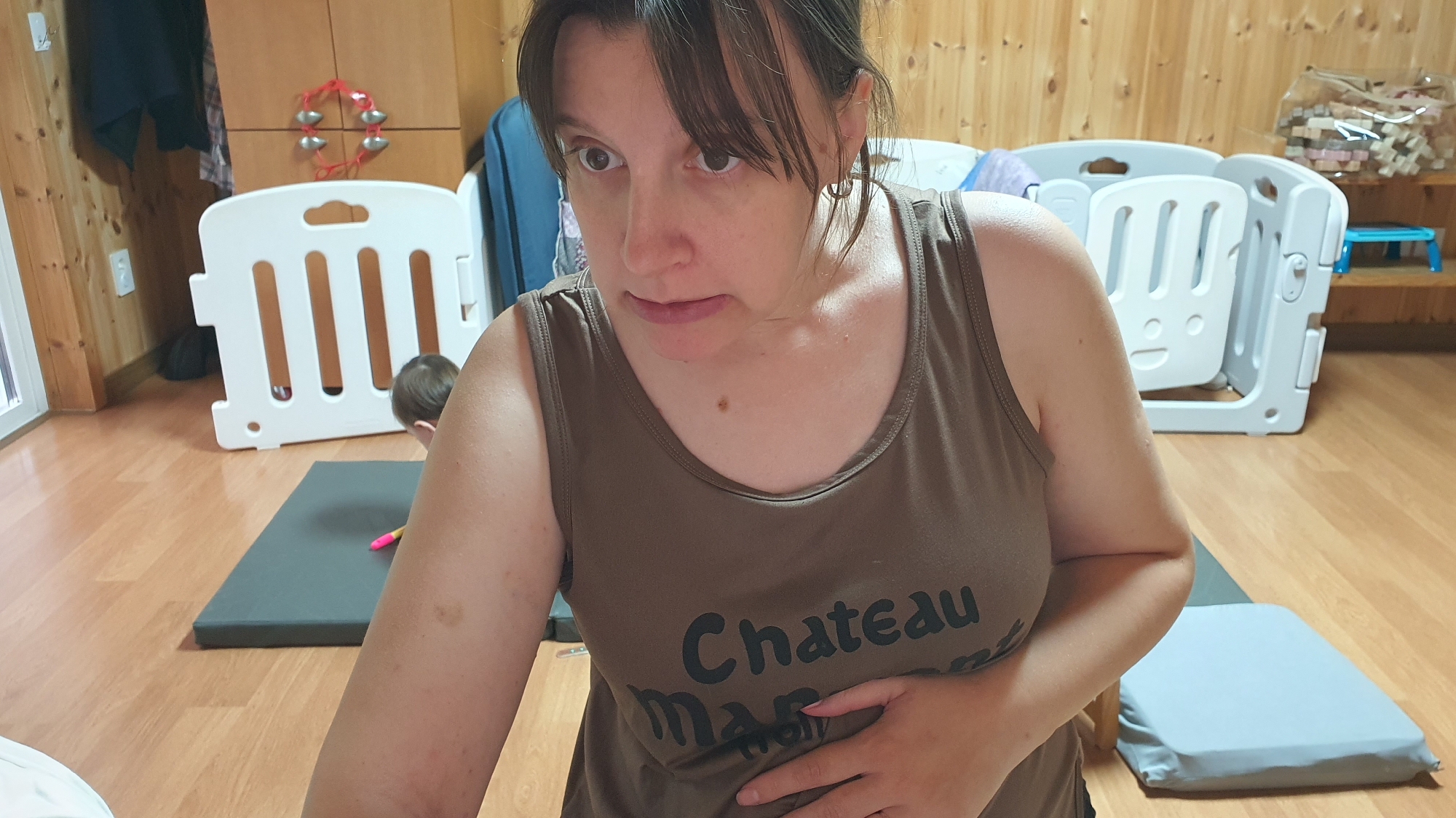 In the original image, a Caucasian woman is positioned in the foreground, facing left and slightly downwards, with her right hand resting on her abdomen. She is wearing a brown tank top with "Chateau Marmont" printed on it in black lettering. Her hair is dark brown and falls to her shoulders. To the left of the woman, a young child with dark hair is visible, facing away from the camera and towards a gray mat on the floor. Behind the child and to the left of the woman is a white plastic playpen. In the background, wooden walls and cabinets are visible, along with some children's toys and a light blue stool. The scene takes place indoors on a wooden floor.