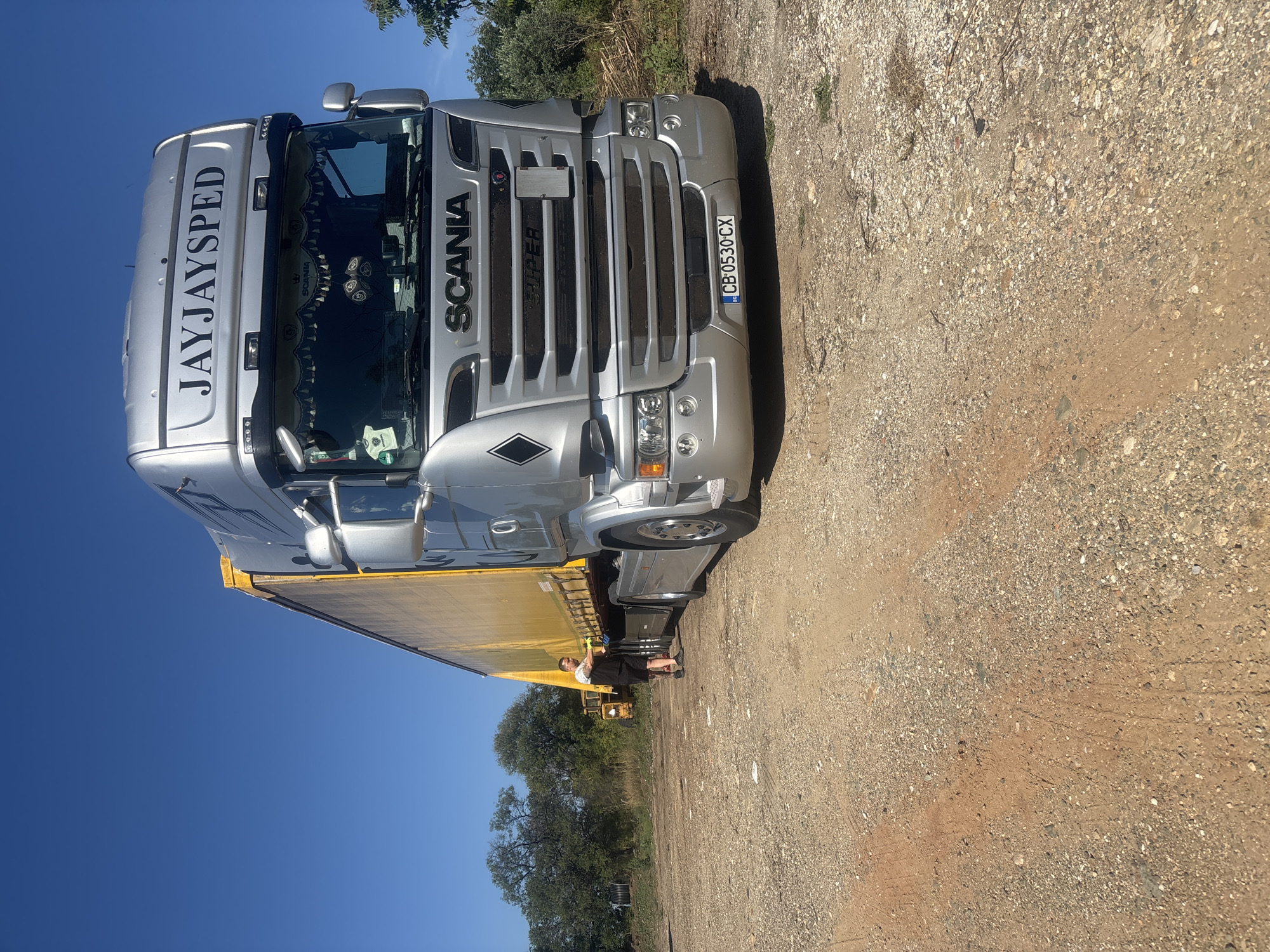 A silver Scania truck with a yellow trailer is parked on a gravel and dirt lot under a clear blue sky. The truck has "JAYJAYSPED" written on the top of its cabin and "SCANIA SUPER" on the front grille. A license plate with "BG 0530 CX" is visible on the front bumper. A man wearing a black t-shirt and shorts is standing near the trailer, appearing to be in the process of securing or checking it. In the background, trees and foliage are visible, along with a yellow construction vehicle. The location is identified as Mikro Doukato, Greece.