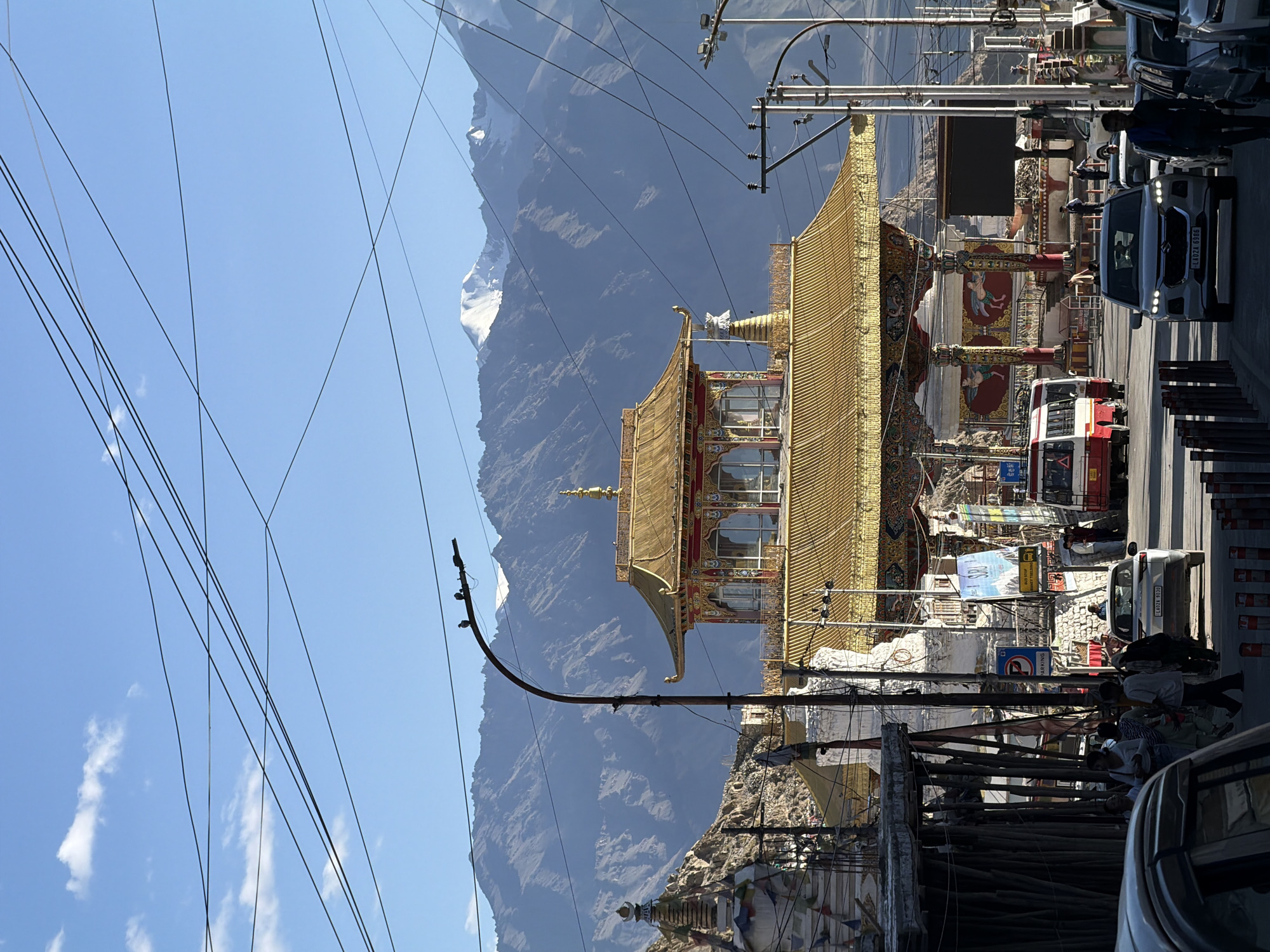 The image captures a street scene in Leh, India, featuring a vibrant golden Buddhist monastery in the foreground, a snow-capped mountain range in the background, and a clear blue sky with scattered clouds. The monastery, with its intricate carvings and traditional architecture, is a prominent focal point. Several cars, a bus, and pedestrians are visible on the street, suggesting a bustling urban environment. The presence of power lines crisscrossing the sky and a street lamp adds to the urban feel of the scene. The overall impression is one of a culturally rich and geographically diverse location, blending religious heritage with everyday life.