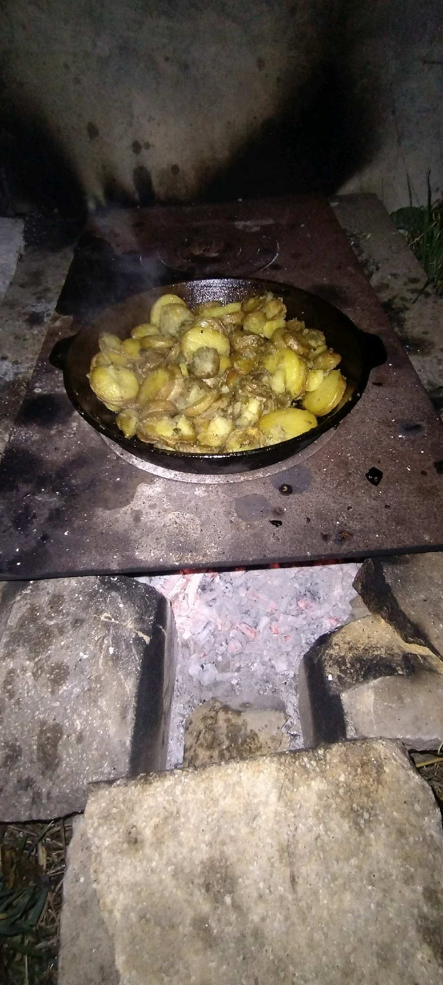 An overhead shot captures a cast-iron skillet filled with sliced potatoes being cooked over a rudimentary outdoor stove constructed from bricks and what appears to be a metal plate. The skillet is placed on the metal plate, which sits above a bed of glowing red embers and white ash. The background shows a textured, dark, and somewhat abstract surface, possibly a wall or a large rock, with shadows cast upon it. The scene is dimly lit, suggesting it is nighttime or dusk, and the overall impression is one of rustic, open-air cooking. The location is identified as Radisne, Ukraine.