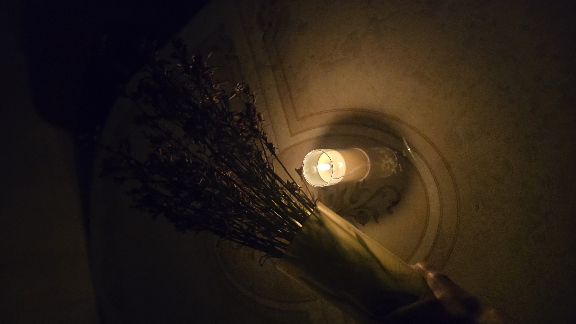 The image depicts a dimly lit scene on a table with a patterned surface. A bouquet of dried, dark-colored flowers, wrapped in a light-colored material, is held by a hand on the left side of the frame. To the right of the flowers, a lit candle in a clear glass holder casts a warm glow. The overall mood is quiet and atmospheric. The location is not specified as Chaikovsky, Russia.