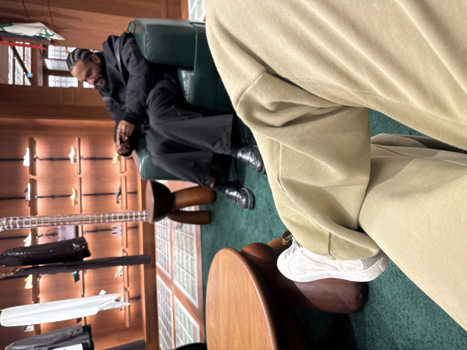 The image depicts an indoor scene within a luxury retail boutique, identifiable as an Hermès store, likely located in Paris, France, given its distinctive design elements such as the specific style of wooden furniture and shelving. The perspective is from a seated individual whose crossed legs, clad in light beige relaxed-fit trousers and a white sneaker with a metallic accent, occupy the lower foreground. A round, dark-stained wooden coffee table is partially visible to the left. In the mid-ground, a male individual with dark skin, a beard, and cornrow braids is seated in a dark green leather armchair. He is dressed in a black puffer jacket, black wide-leg trousers, and black loafers. He is observed with a slight smile, looking towards his right, with his hands positioned near his lap. Between the foreground and mid-ground subjects, a unique three-legged wooden stool with a dark circular top and a clear, ridged display column are present. The background features warm-toned wooden wall shelving units with integrated lighting, showcasing various luxury shoes and a purple handbag. Garments, including a white piece and a dark item, hang on a rack to the far left. The floor of the immediate seating area is covered by a deep green carpet, transitioning to a patterned floor with light square tiles in the distant background.