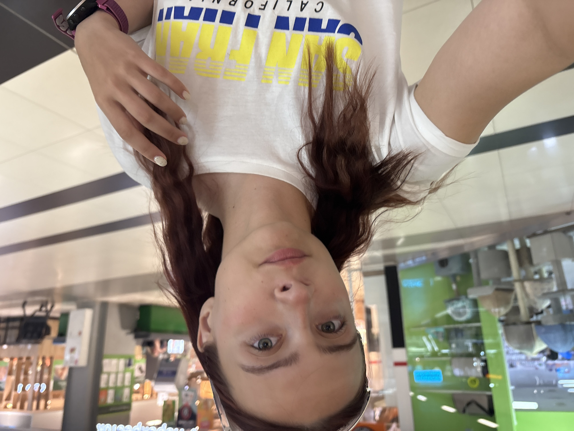 A young woman with long, dark-brown hair and green eyes is taking a selfie in a brightly lit indoor commercial space. She is wearing a white t-shirt with yellow and blue lettering that reads "CALIFORNIA". A pink smartwatch is visible on her left wrist. Her nails are painted a light, neutral color with a few subtle dots. The background appears to be a retail environment, with shelves of products and tiled flooring. The image is oriented vertically, with the woman's face closest to the camera.
