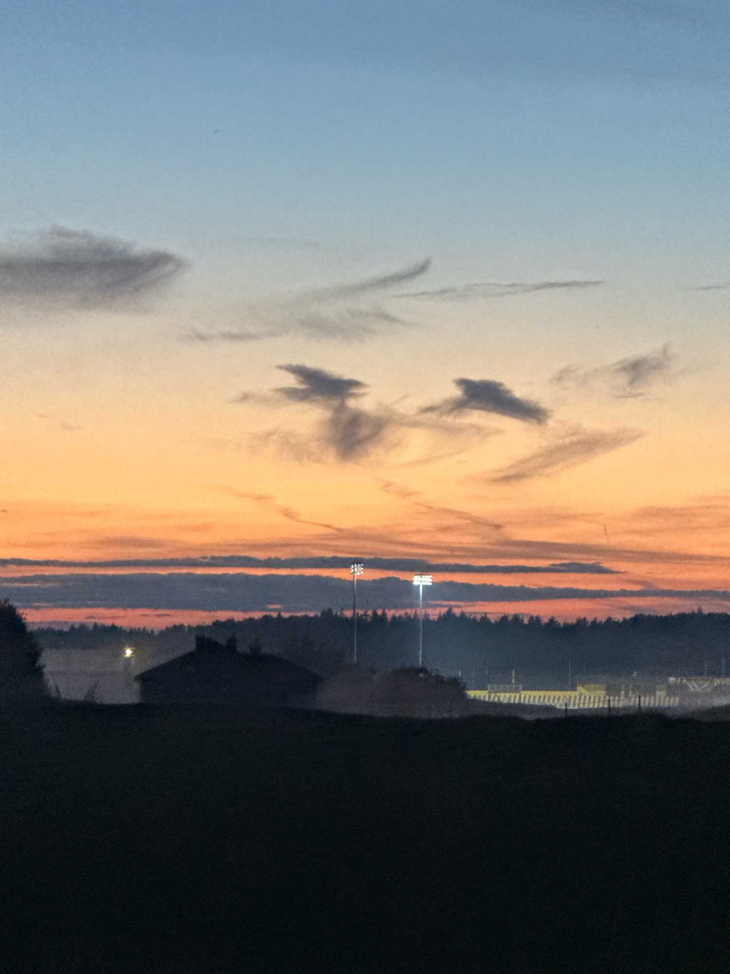 The image depicts a twilight scene over what appears to be a rural area on the outskirts of Galinė, Lithuania. The sky transitions from a pale blue at the top to a vibrant orange and then a deep pinkish-red towards the horizon. wispy, dark clouds are scattered across the sky.
In the middle ground, a dark silhouette of a building with a pitched roof is visible, possibly a residential home, with a faint light emanating from its vicinity. To the right of this building, a line of trees forms a dark band across the landscape. Beyond the trees, a row of tall, illuminated stadium lights is prominent, suggesting a sports field or stadium. In the far distance, a fence-like structure with a yellow top is faintly discernible. The foreground is a dark, out-of-focus field. The overall impression is of an early morning or late evening in a quiet, possibly agricultural or recreational area outside of a populated center.