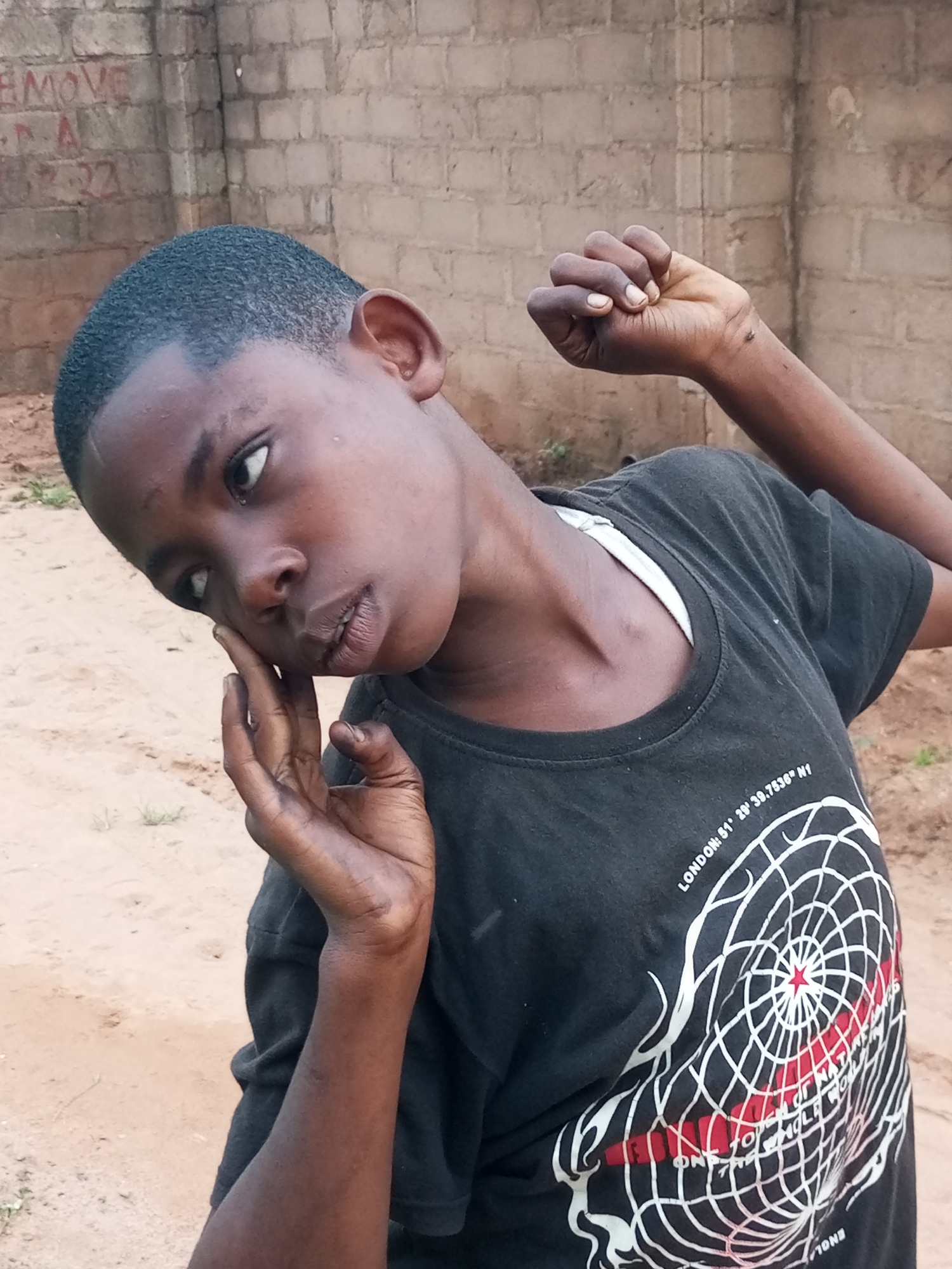 A young African boy is captured in a close-up portrait, his face turned away from the camera, looking to his left with a thoughtful or questioning expression. His right hand is raised and touches his cheek and temple, with his fingers slightly splayed, while his left arm is bent at the elbow with his fist clenched and raised. He is wearing a black t-shirt with a white undershirt visible at the neckline. The t-shirt features a graphic design of a spiderweb-like pattern with a red star in the center and some text.
The background suggests an outdoor, possibly rural or semi-urban setting. A brick wall, some of which has graffiti, forms the backdrop. The ground appears to be dusty or sandy, with some sparse vegetation visible. The lighting is natural, indicating it is daytime. The image's focus is on the boy, with the background being somewhat blurred, drawing attention to his pose and facial expression. The provided information about the location being in Tempure, Nigeria, is external to the visual content of the image itself.