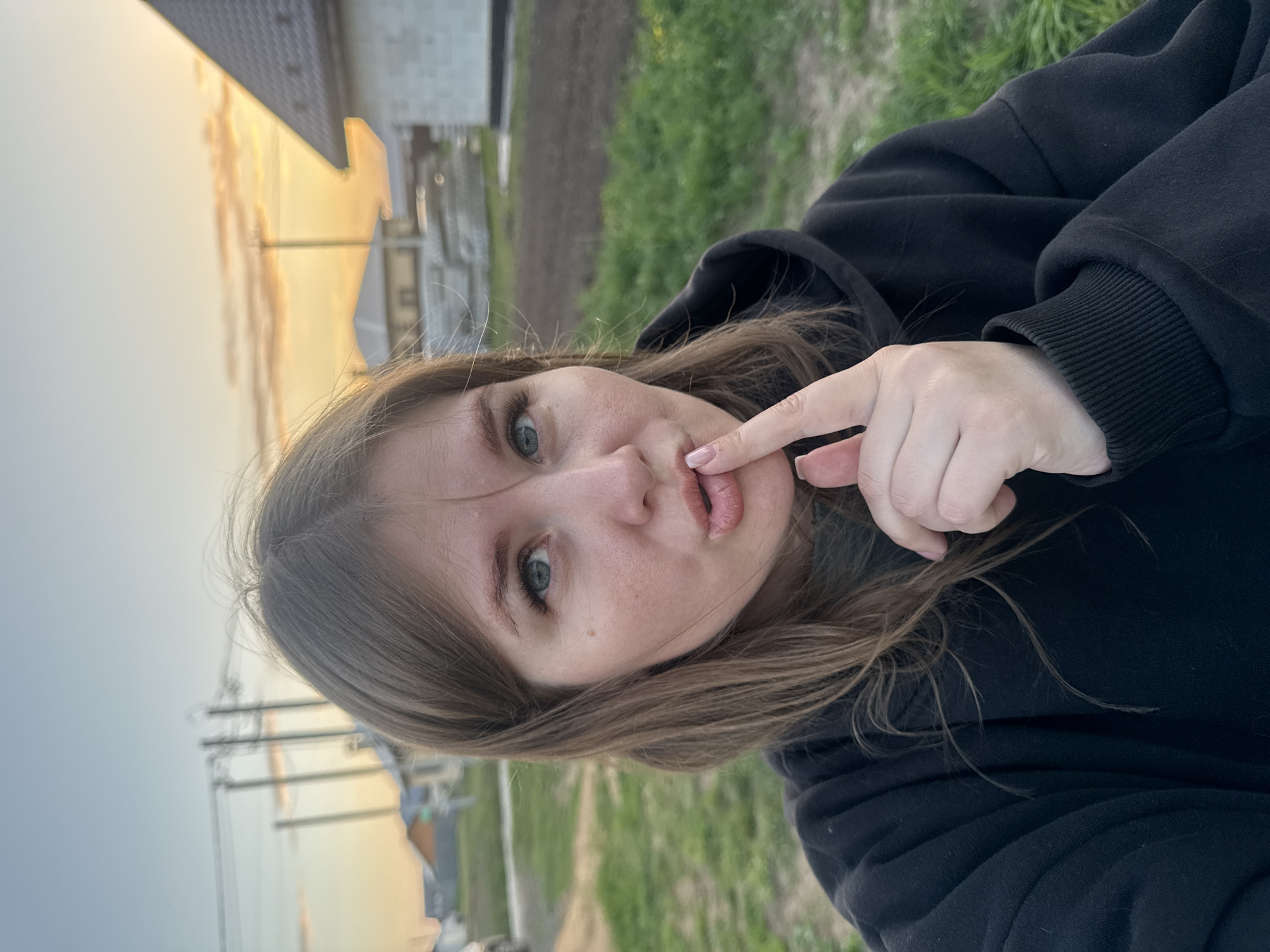 A woman with light brown hair and green eyes is holding her finger to her lips. She is wearing a black hoodie. The background includes a building, utility poles, and a field. The location is Ферма, Russia. The sky is a mix of orange, yellow, and gray.