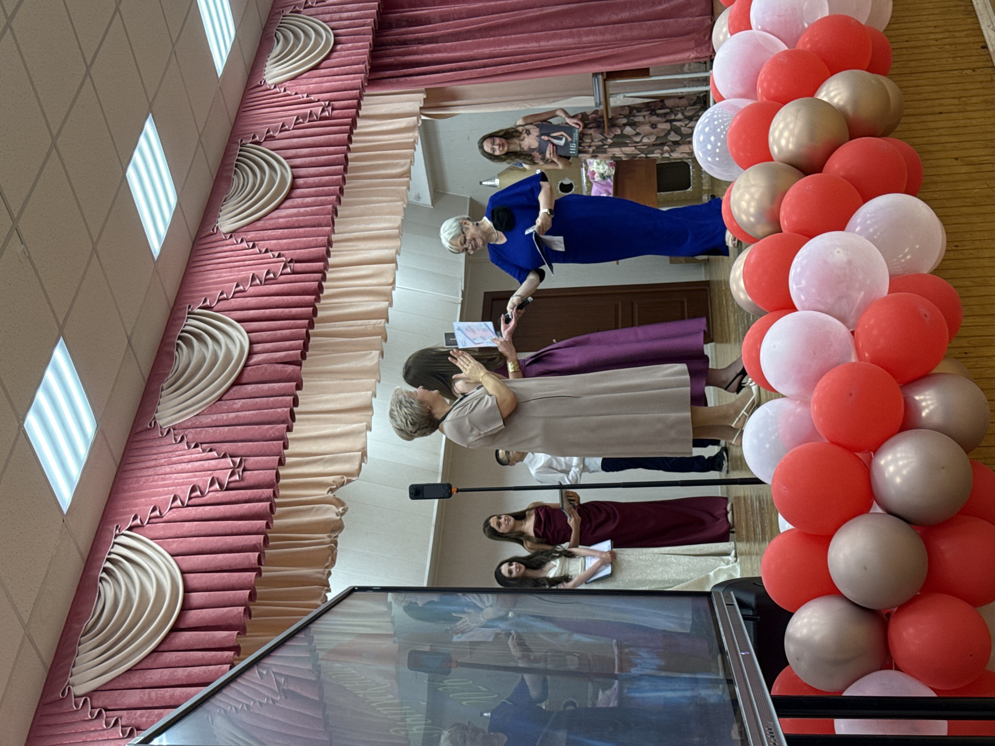 This media file captures a scene at what appears to be an indoor event, possibly a graduation or awards ceremony, given the formal attire of the individuals. The setting includes decorative elements such as pleated fabric draping in shades of pink and beige, and a balloon arch composed of red, white, and gold balloons. The lighting suggests an indoor environment, with fluorescent ceiling lights visible at the top.
Several people are present. In the foreground, a woman in a blue dress is holding a microphone and speaking, facing a group of individuals. To her left, another woman in a purple dress is partially visible. Further back, a young woman in a dark red dress is holding a magazine, and another young woman in a cream-colored dress is also present, both looking towards the speaker. A large screen or display is also visible, reflecting some of the scene.
The overall atmosphere seems formal and celebratory. The people's expressions are largely unreadable due to the image orientation and resolution, but the event appears to be well-attended and organized. No specific time of day or weather conditions can be determined from the image. Location cues point to Moscow, Russia, but there are no explicit signs or text within the image to confirm this. The visible text is limited to what appears to be the cover of a magazine held by one of the attendees, but the text is too blurry to read.