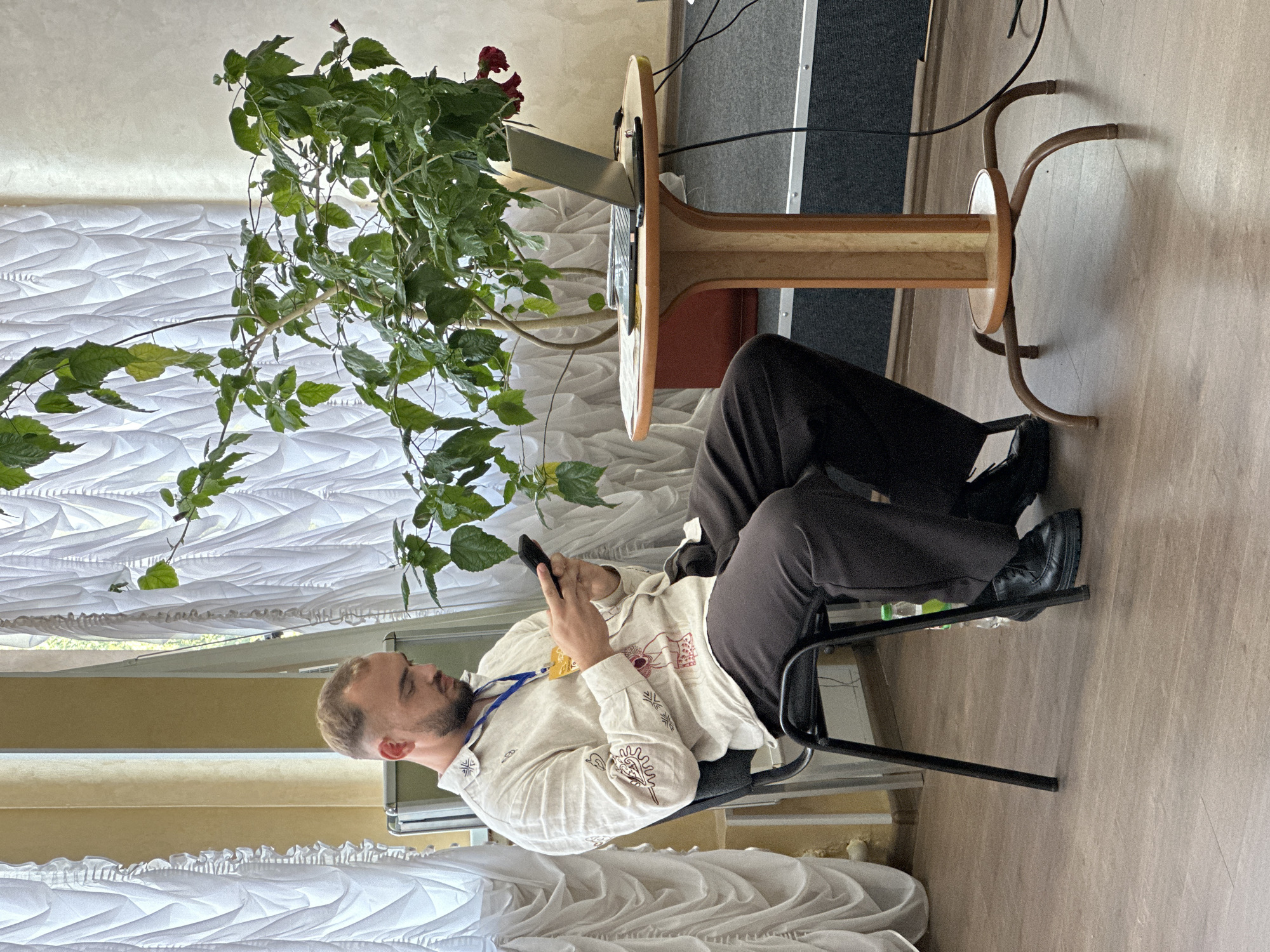 In this media file, a man is seated in a black chair, leaning back and looking down at his phone, which he holds in both hands. He is wearing a light-colored shirt with dark embroidered designs and dark trousers. A blue lanyard with a yellow badge is around his neck. He has a beard and short hair. The scene appears to be indoors, with a wooden floor. To his left, a large potted plant with green leaves and a few red flowers is prominent. Behind him, a round wooden table with a light-colored top and dark metal legs is visible, with a laptop on it. The background shows white curtains and a hint of a window. The overall lighting suggests daytime. The image is oriented vertically, with the man appearing in the lower portion of the frame.