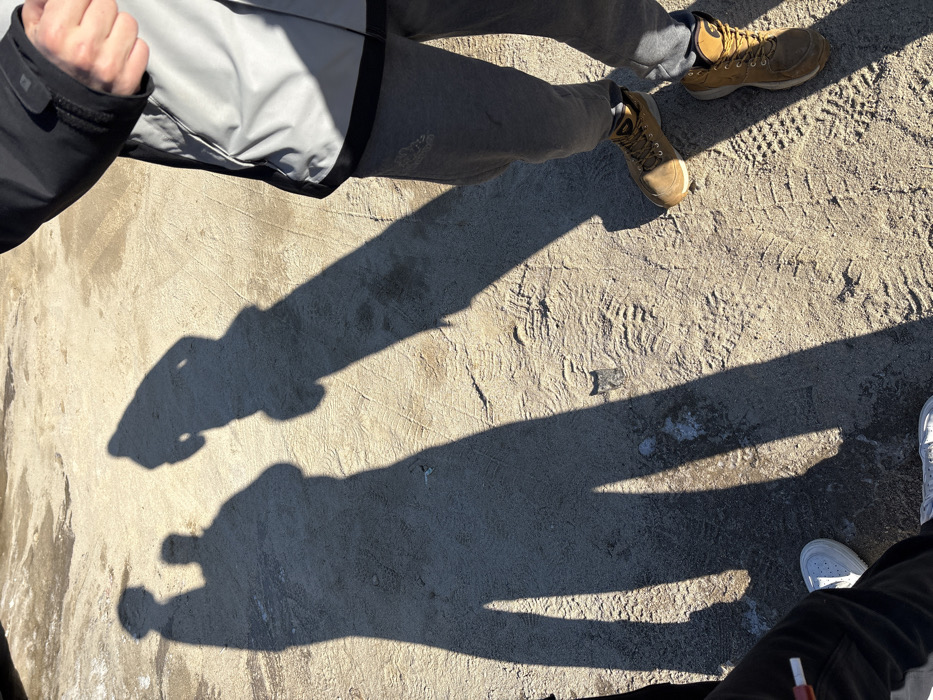 The image displays an outdoor scene in Bistritsa, Bulgaria, featuring a light tan, sandy or dirt ground marked by multiple tire tracks and various boot prints. Several patches of a lighter, possibly icy or frozen, substance are visible on the ground. Direct sunlight from a low angle casts long, distinct shadows of at least three human figures across the terrain.
In the right foreground, the lower torso, legs, and feet of one individual are visible. This person wears a black and light grey two-toned jacket, grey athletic sweatpants with a dark logo partially visible on the left thigh, and tan, high-top hiking boots with dark soles and laces. A partially obscured hand, appearing clenched, is visible near the jacket's right sleeve.
In the lower-left foreground, the lower legs and feet of a second individual are present. This person wears dark trousers and white athletic sneakers with white laces. A white, cylindrical object, resembling a cigarette or vape pen, extends from the area near their left leg.
Three elongated human shadows are cast prominently across the ground. One shadow corresponds to the person in the right foreground. Two additional shadows are located in the center-left, standing upright and appearing to be of two individuals. The shadow on the far left is partially cropped. No direct interaction between the visible individuals or their shadows is discernible beyond their proximity.