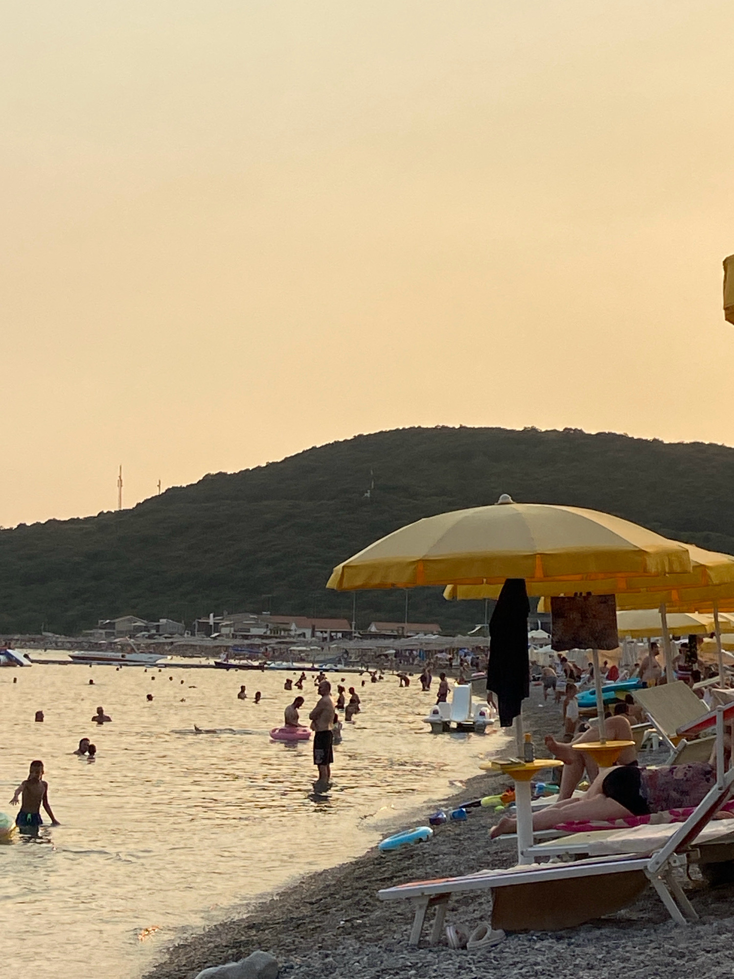 The image depicts a crowded beach scene on a clear day with a hazy, pale orange sky. In the foreground, a beach with pebbles and sand is visible, with several lounge chairs and yellow beach umbrellas. One lounge chair in the foreground has a person lying on it. The water is calm and shallow, with numerous people swimming, wading, and playing. Some individuals are on inflatable toys. In the middle ground, there is a line of buildings and more beach umbrellas. In the background, a large, green, tree-covered hill dominates the landscape. The scene appears to be during daylight hours, possibly late afternoon or early evening due to the sky's color. The location is identified as Prijevor, Montenegro.