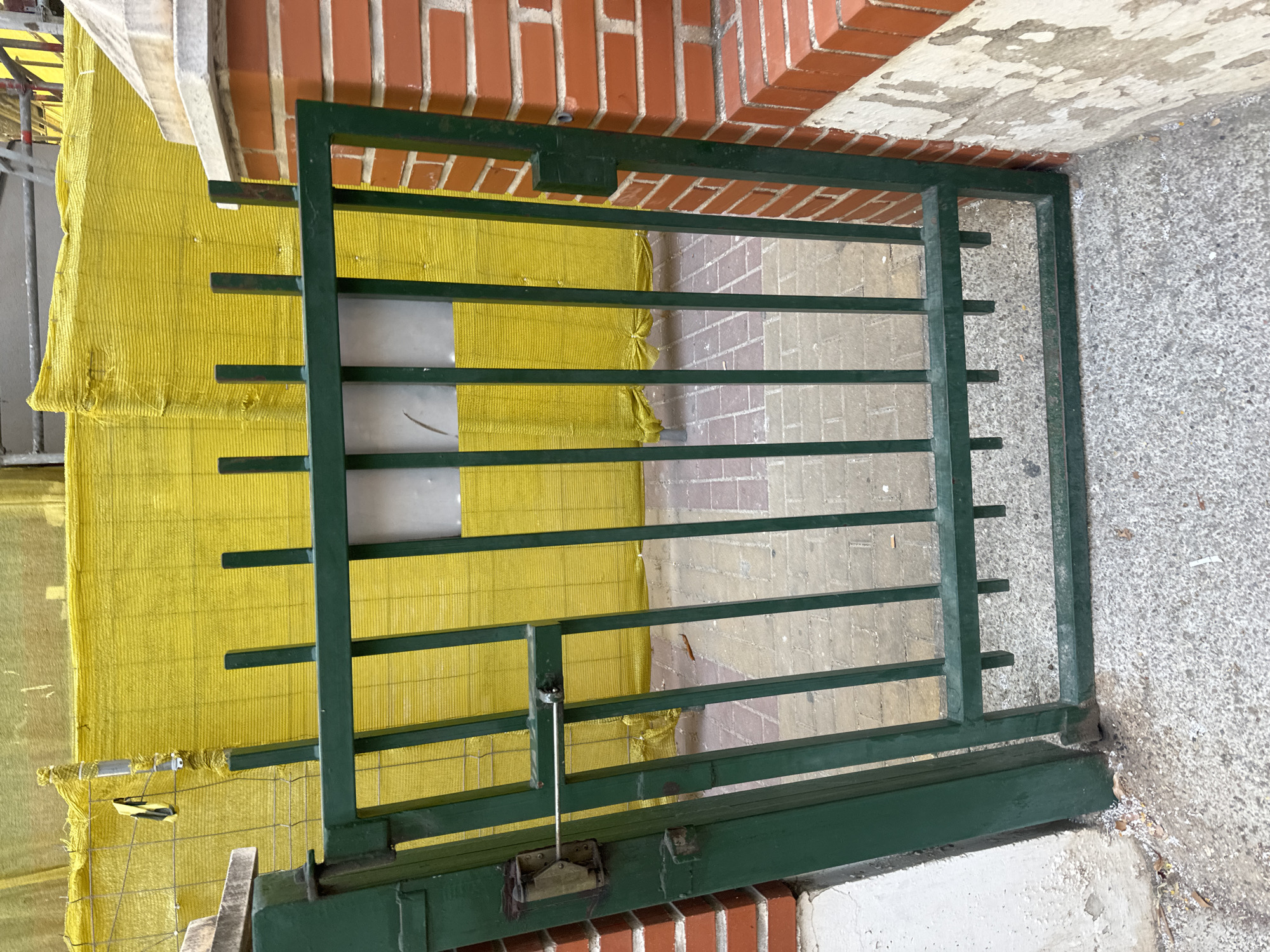 The image shows a green metal gate in front of a yellow construction barrier. The gate is closed and has vertical bars. To the right of the gate, there is a brick wall with a concrete cap. The ground in front of the gate is paved with concrete and cobblestones. The scene appears to be an exterior shot, possibly in a city environment. The yellow barrier suggests ongoing construction or maintenance work. The location is identified as Murcia, Spain. There are no people or animals visible in the image.