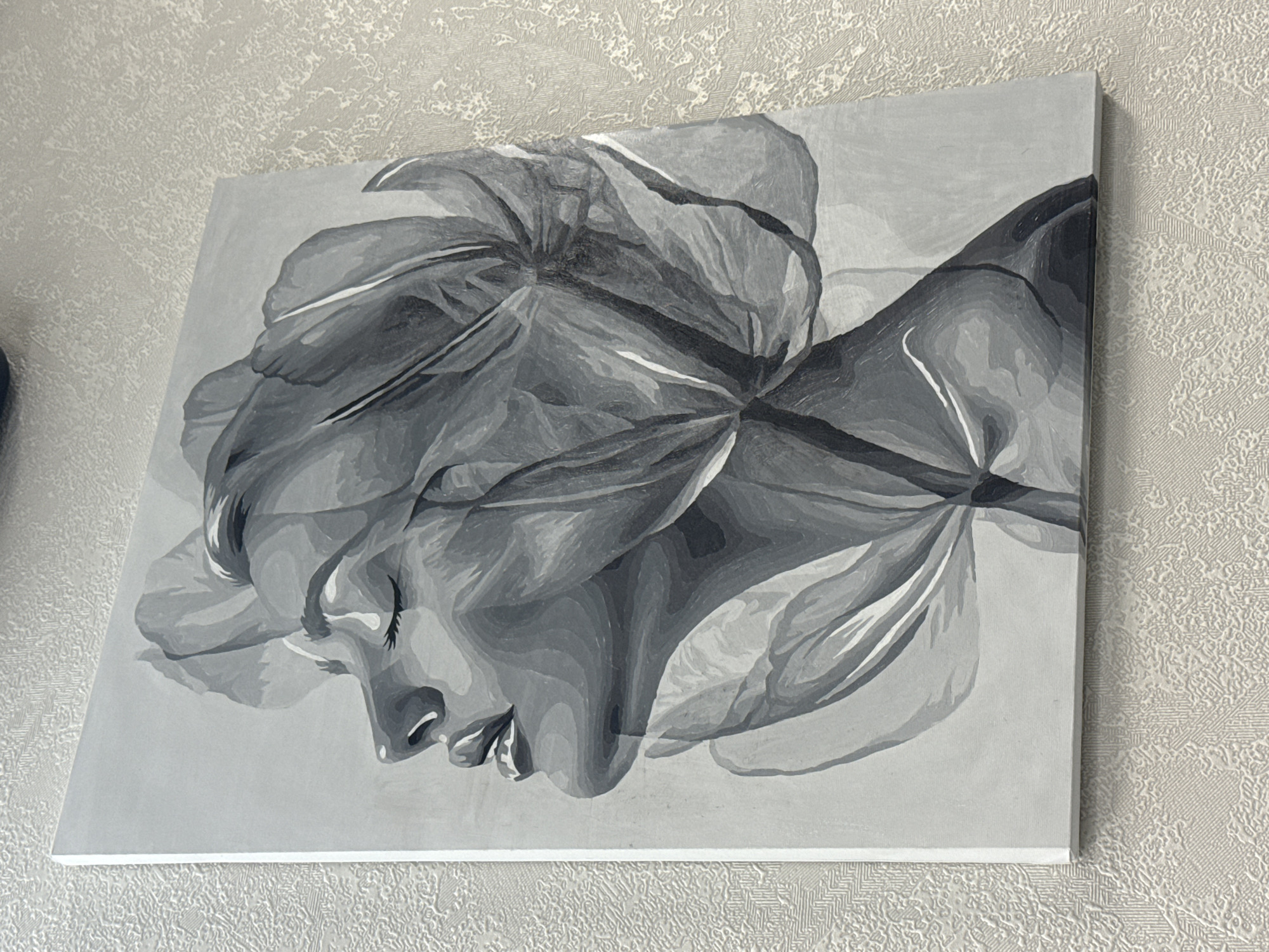 The image shows a black and white painting of a woman's face interwoven with the petals of a flower. The painting is mounted on a wall with a textured, patterned wallpaper. There are no visible people, objects, or interactions besides the artwork itself. The location cannot be determined from the image alone.