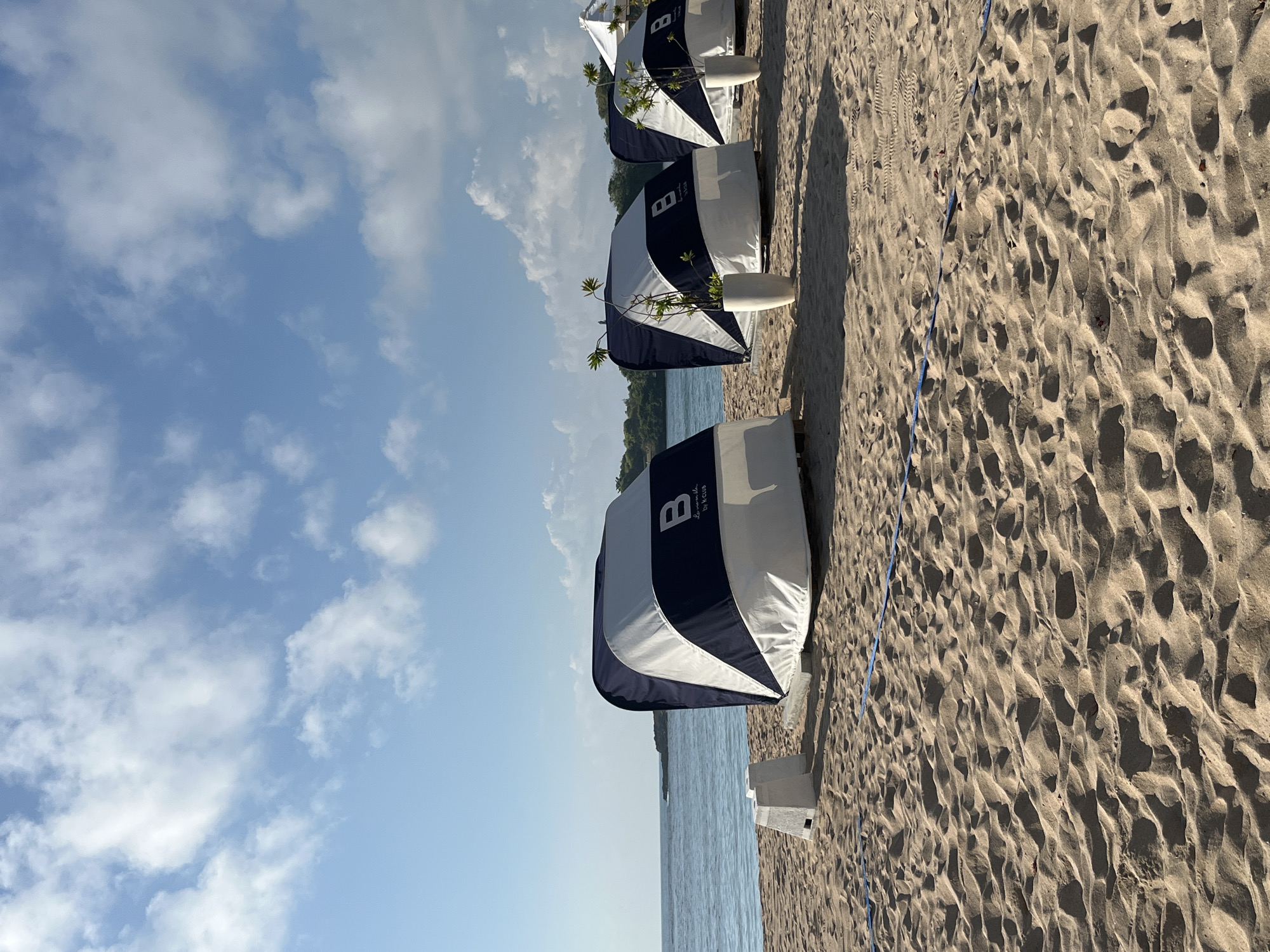 A view of several cabanas on a sandy beach in Kuta Selatan, Indonesia. The cabanas are blue and white with a "B by K Club" logo. They are set up in a row facing the ocean, which is visible in the background. The sky is blue with scattered white clouds. The sand is light brown and appears textured with footprints and ripples. In front of the cabanas, there are some white planters with greenery. A blue line is visible on the sand.