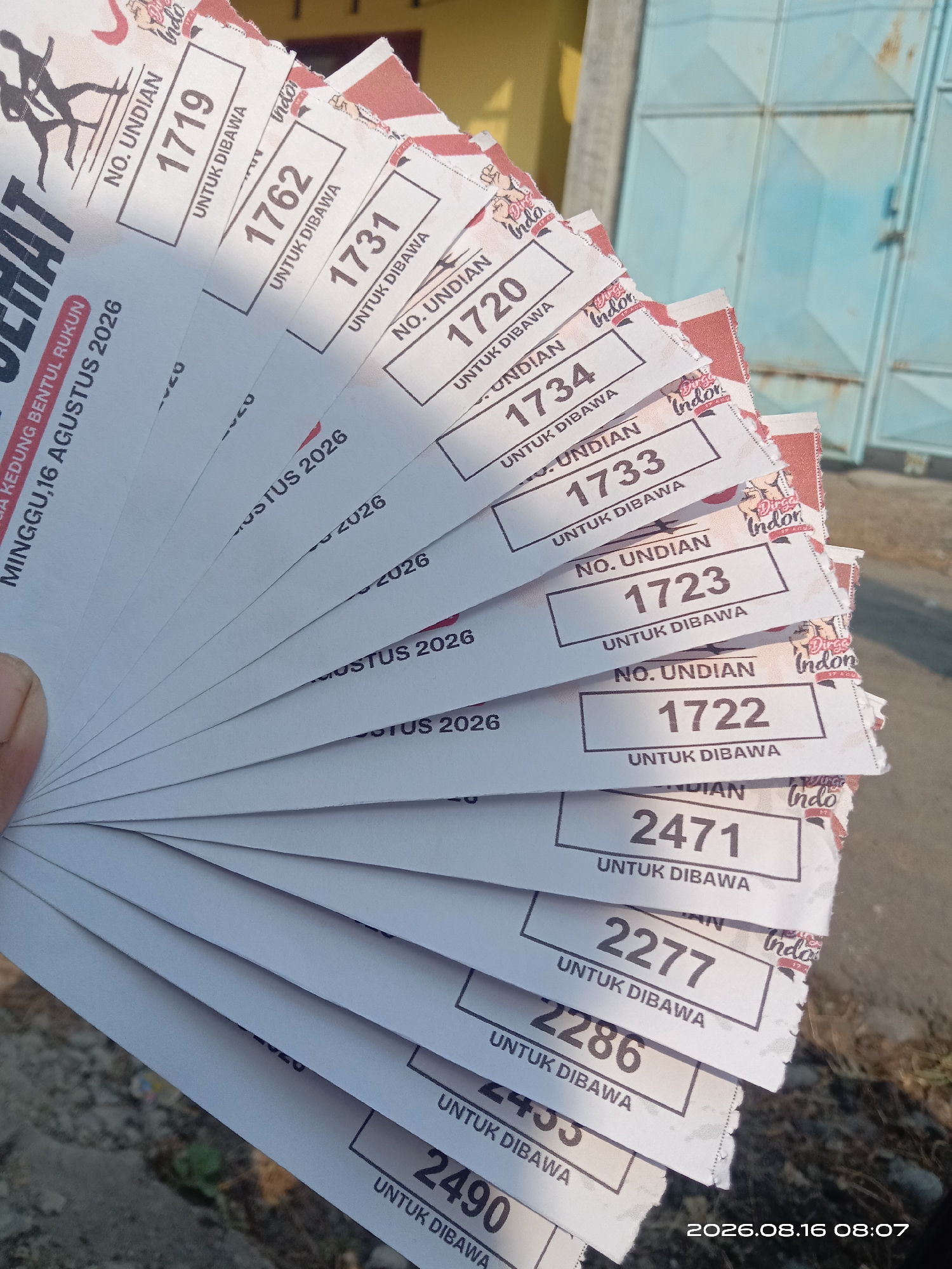 The image shows a fan of raffle tickets held in a person's left hand. The tickets are white with red and black printing. The top ticket visible has the number 1719. Subsequent tickets are numbered 1762, 1731, 1720, 1734, 1733, 1723, 1722, 2471, 2277, 2286, 2453, and 2490. Some tickets also have text that reads "NO. UNDIAN", "UNTUK DIBAWA", and "MINGGU, 16 AGUSTUS 2026". The background appears to be an outdoor setting with a blue metal door or gate and a building with a yellow wall. The ground is visible at the bottom of the image and appears to be a dirt or gravel surface. A date stamp in the lower right corner reads "2026.08.16 08:07". The overall impression is of a close-up shot of raffle tickets being displayed outdoors. The mention of "Banyuarang, Indonesia" is not directly supported by visual information in the image, but the text on the ticket mentioning "MINGGU, 16 AGUSTUS 2026" suggests an Indonesian context.