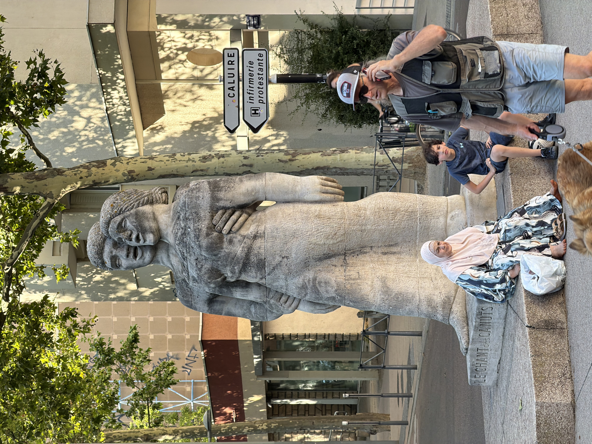 This media file captures an outdoor scene in Lyon, France, featuring a large stone statue and several people. The statue, an abstract representation of two figures embracing, dominates the foreground. A man, wearing a vest and a baseball cap, is seen holding a phone to his ear while also appearing to film. He is standing near a young boy who is sitting on the edge of a concrete ledge, looking towards the camera. Beside the boy, a woman wearing a headscarf and a patterned dress is also seated on the ledge, her feet bare. A fluffy dog, partially visible at the right edge of the frame, appears to be on a leash held by the man.
The scene is bathed in sunlight, creating dappled shadows on the ground and a building in the background, suggesting it is daytime. The presence of trees and the architectural style of the building indicate an urban environment. Signage is visible in the background, with one sign reading "CALUIRE" and another with "H infirmerie protestante," providing specific location cues within Lyon. A partially visible inscription on the base of the statue reads "LE CHANT des CANUTS." The overall mood appears casual and observational, with the people seemingly engaged in their own activities or interactions.