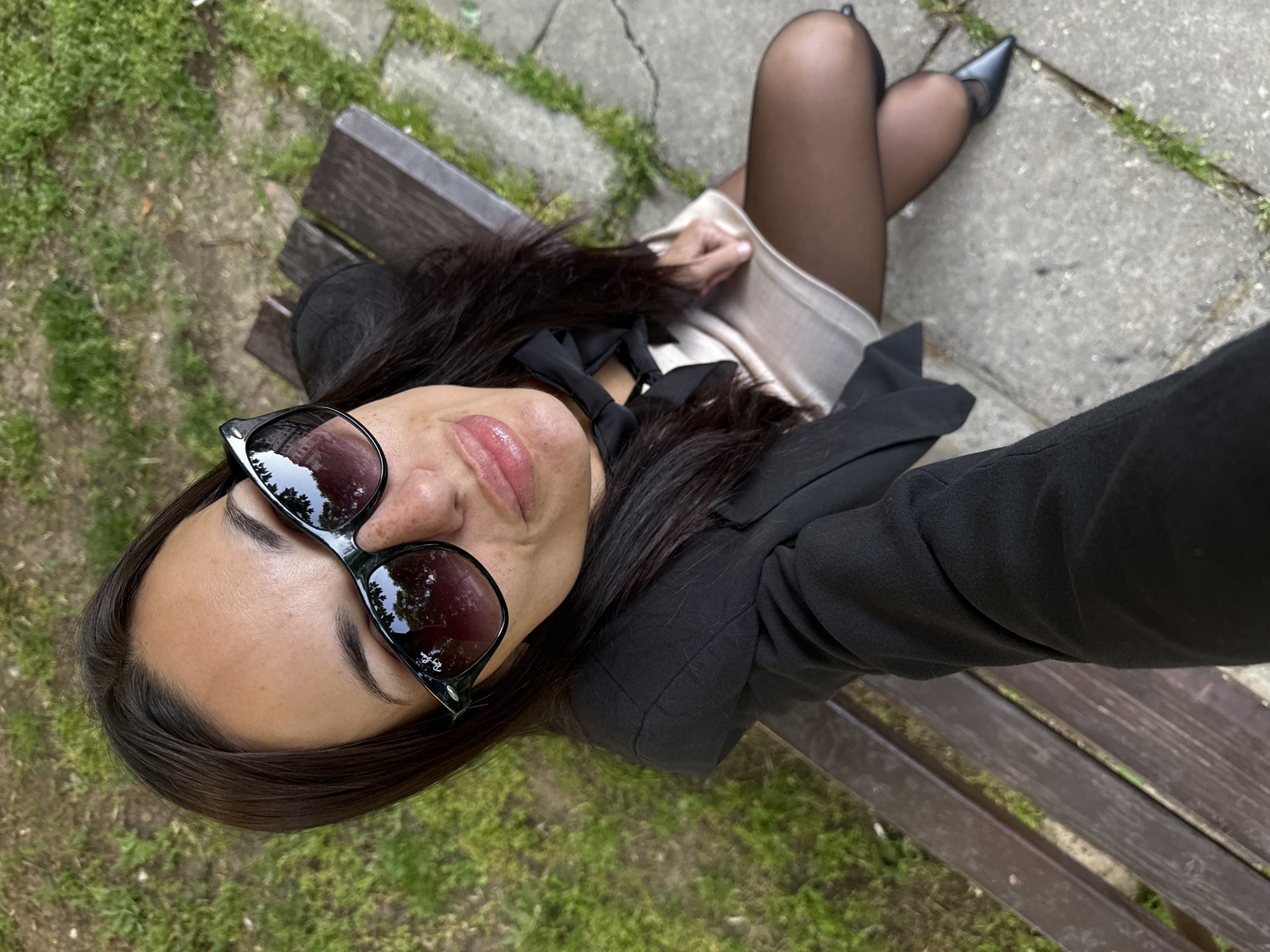 A young woman wearing a black blazer, light-colored dress, black tights, and black pointed-toe heels is reclining on a wooden bench, taking a selfie. She is also wearing sunglasses and a black bow around her neck. The bench sits on a paved surface with grass growing in the cracks. Based on the reflections in the sunglasses, she may be in a park or other green space. The location is in Pleven, Bulgaria, as evidenced by the image context.
