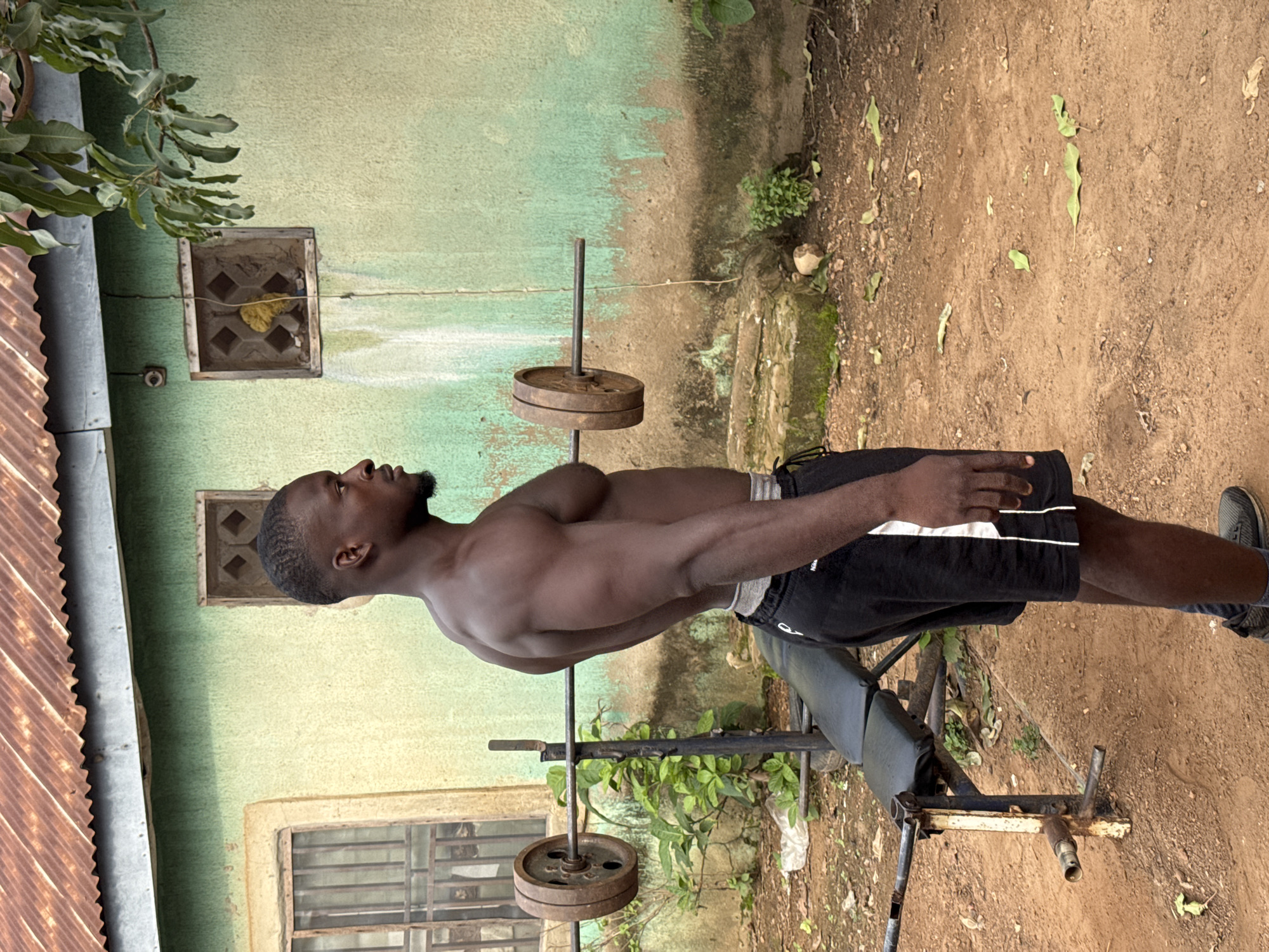 The image, originally rotated 90 degrees clockwise, displays a shirtless young adult male standing outdoors next to weightlifting equipment. The man, with dark skin, a short beard, and a muscular physique, is wearing black athletic shorts with white trim. He is positioned in profile, looking upwards and to his left, suggesting he is either posing or taking a break from a workout.
Next to him is a simple, black padded weight bench with a metal frame, and a metal barbell stand holding a barbell loaded with several rusty, round weight plates. The setting is an unpaved outdoor area with reddish-brown dirt ground, scattered with dry leaves and small green plants.
In the background, a light green building wall features two small, square window openings; one contains a decorative concrete block pattern. To the far left, rusty corrugated metal sheeting, possibly a roof or fence, is visible. Green leaves and branches from a tree extend into the frame from the top left. A thin wire also runs horizontally across the upper middle ground.
The scene depicts daytime with bright, natural light, indicating clear weather. The rustic nature of the equipment and the outdoor setting suggest a home gym in a rural or semi-urban environment, fitting the Akwanga, Nigeria context. The man's expression is neutral. No text is visible in the image.