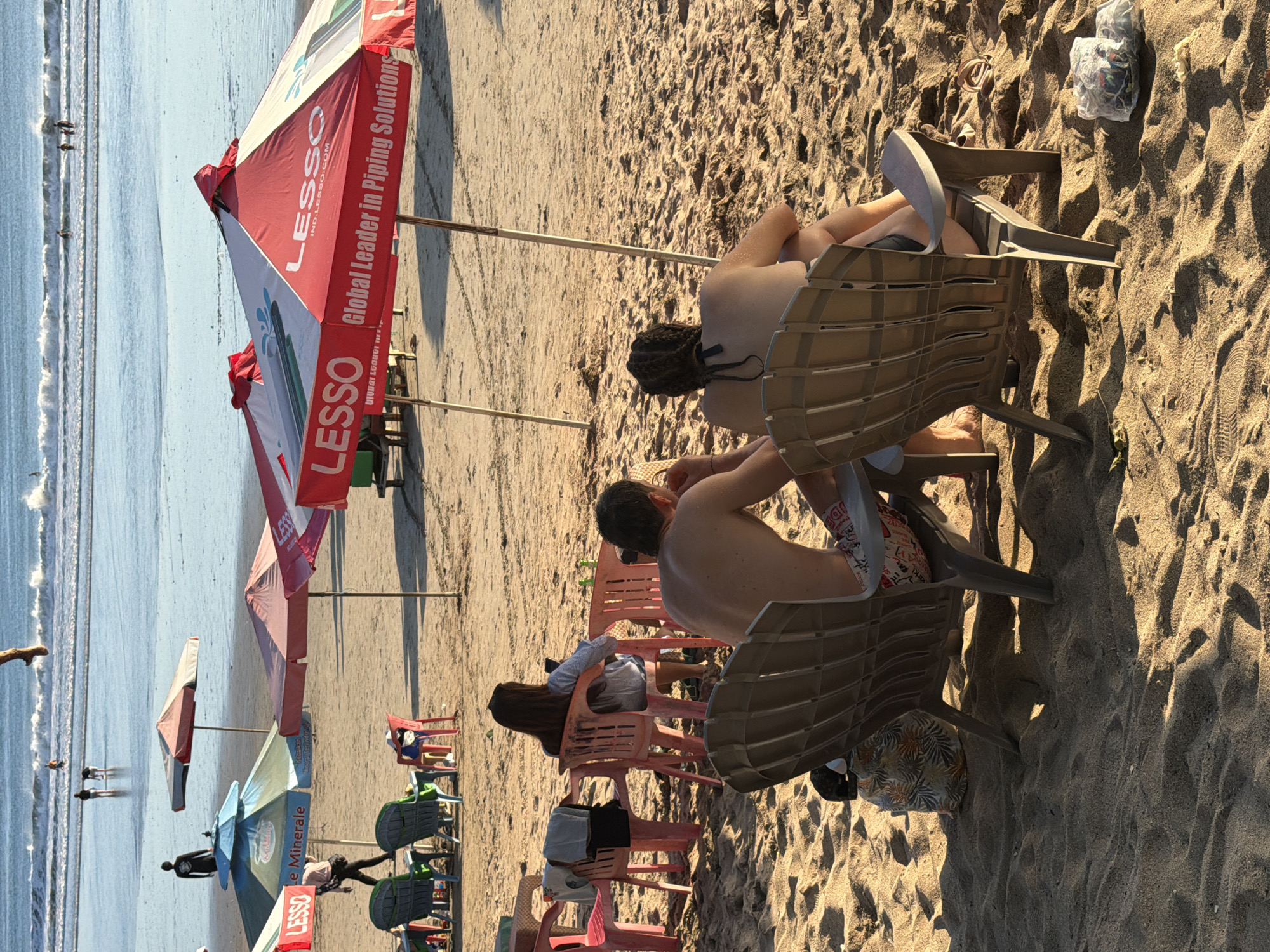 This is a photograph taken on the beach in Kuta, Indonesia, during the late afternoon. In the foreground, two individuals are seated in beach chairs facing away from the camera. The person on the left appears to be a man with bare shoulders, wearing patterned shorts. The person on the right, also with bare shoulders, is a woman with braided hair. In the middle ground, a woman with long dark hair is seated on a pink plastic chair, looking at a mobile phone. Behind her, a man in dark clothing stands facing the ocean. Several beach umbrellas are visible, with the prominent red and white "LESSO" umbrella featuring text that reads "Global Leader in Piping Solutions." Other colorful umbrellas and empty plastic chairs are scattered across the sandy beach. In the background, the ocean waves are breaking, and several people are in the water. The lighting suggests the sun is low in the sky, casting long shadows. A patterned bag rests on the sand next to the left beach chair, and a plastic bag containing items is on the sand in the lower right corner.
