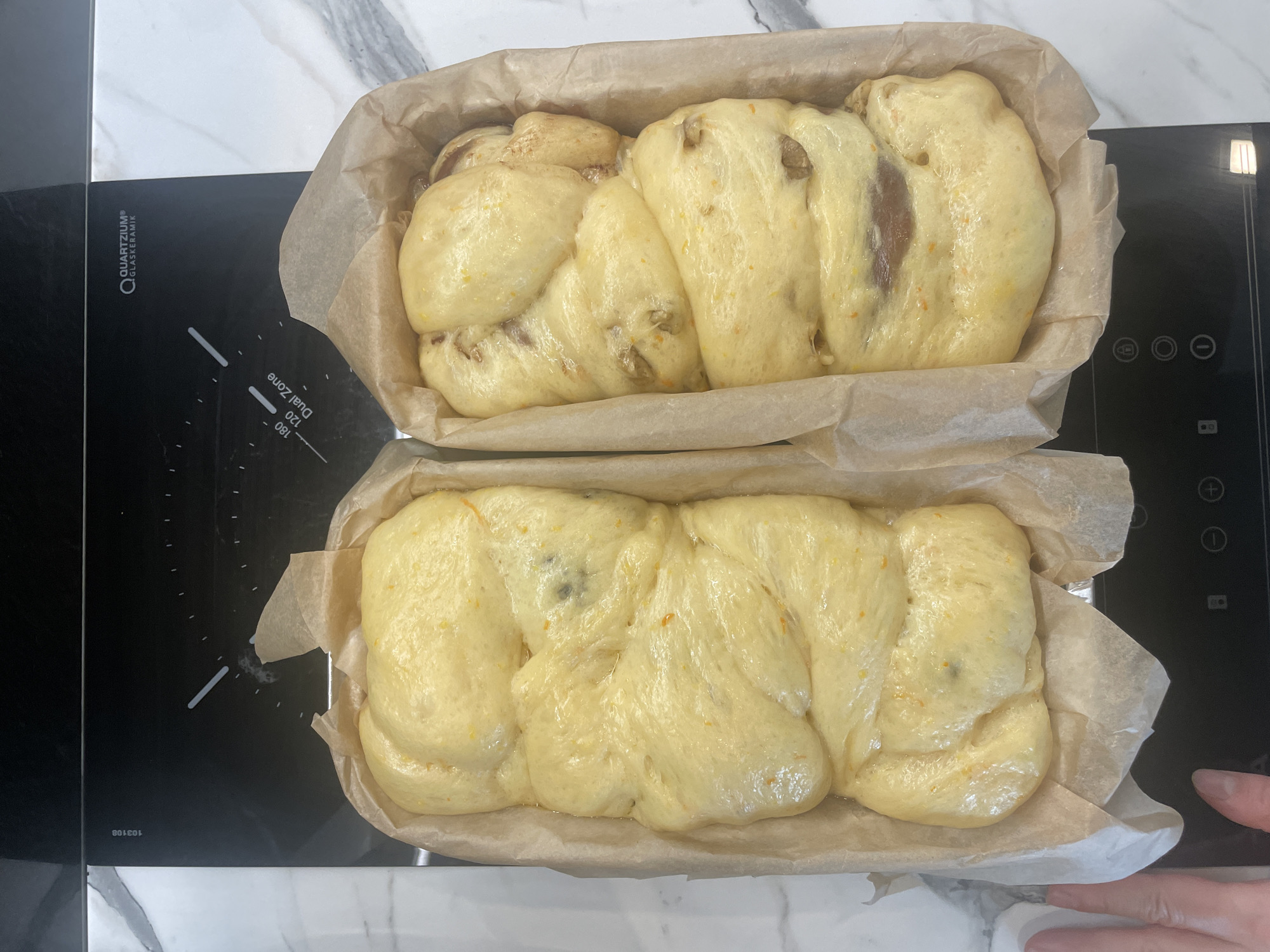 Two loaf pans lined with parchment paper, each containing a braided bread dough, rest on a black induction cooktop. The dough in the top pan appears to have visible additions such as nuts and dark fruit, while the bottom loaf appears to only have citrus zest. The cooktop is situated atop a white countertop with marble patterning. The image was likely taken indoors. While the exact location in Sofia, Bulgaria is not determinable from the image, the cooktop is positioned on a countertop typical of a modern kitchen. A finger from a human hand is visible in the bottom right corner of the image.