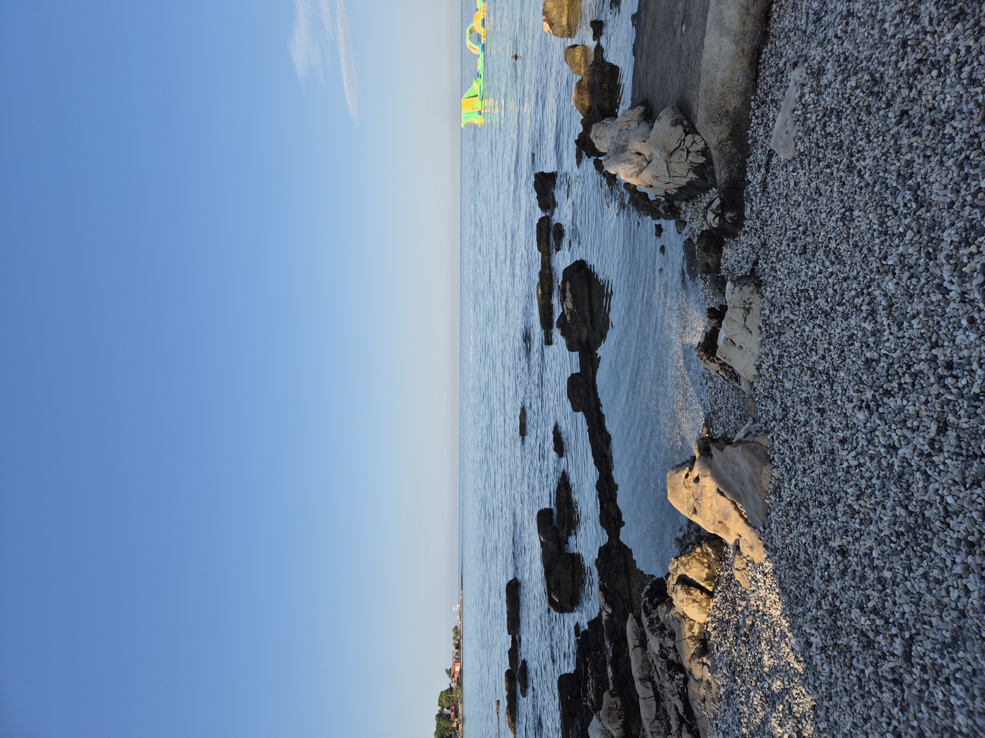 The image depicts a rocky shoreline on a clear day in Monterol, Croatia. The foreground is composed of a dense layer of small, light-colored pebbles, interspersed with larger, sun-drenched rocks. In the mid-ground, dark, jagged rocks protrude from the calm, blue water of the Adriatic Sea. The water reflects the clear sky, which occupies the upper half of the frame. In the distance, a thin line of trees and buildings, likely hotels or apartments, can be seen along the coast. Further out in the sea, colorful inflatable water toys, a green and yellow structure with an arched section, are visible. A small, indistinct human figure can be seen near the inflatable toys. The overall scene suggests a tranquil beach environment, potentially during the daytime.